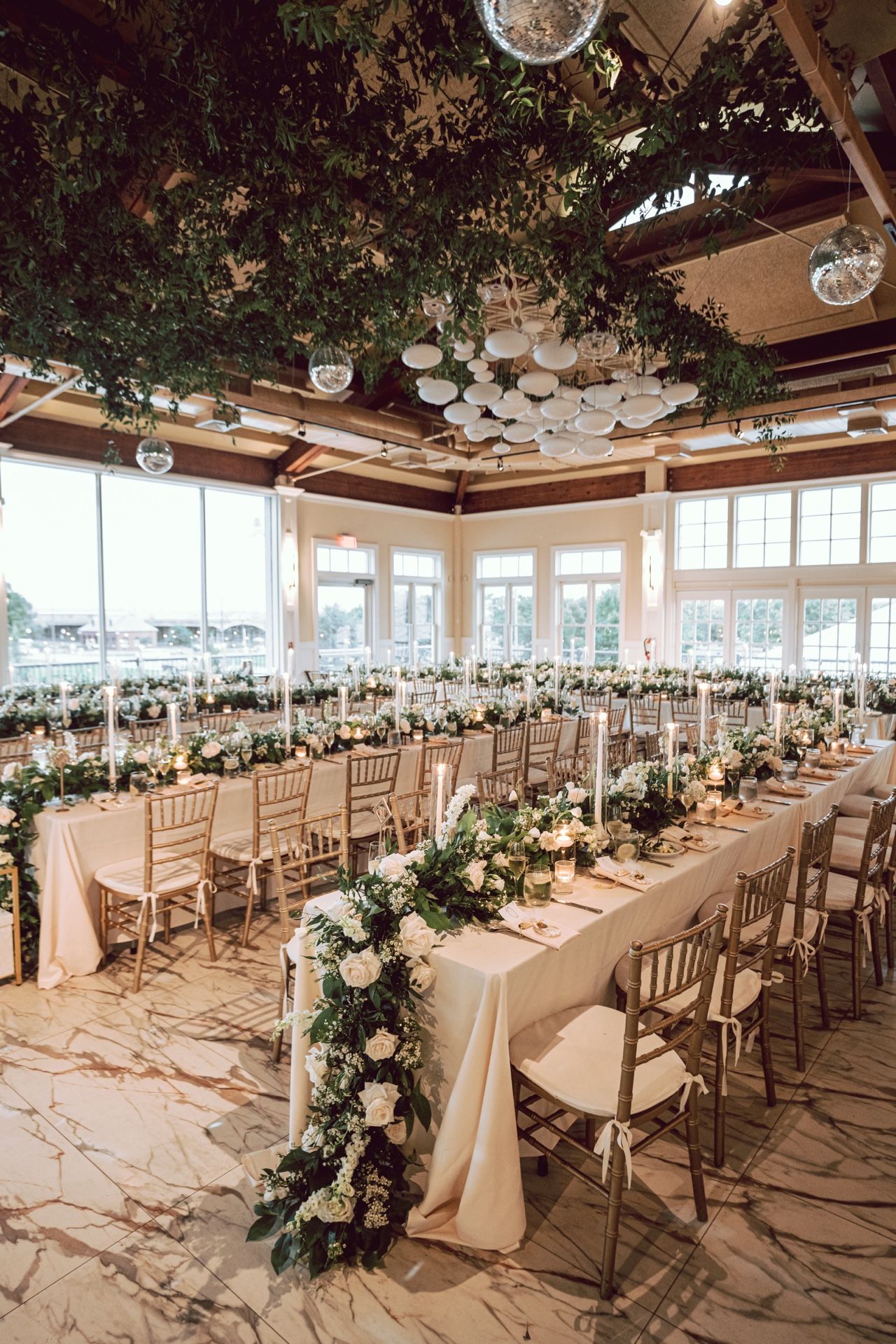 Elegant wedding reception hall with floral garlands, marble floors, gold chiavari chairs, and lush greenery ceiling decor.