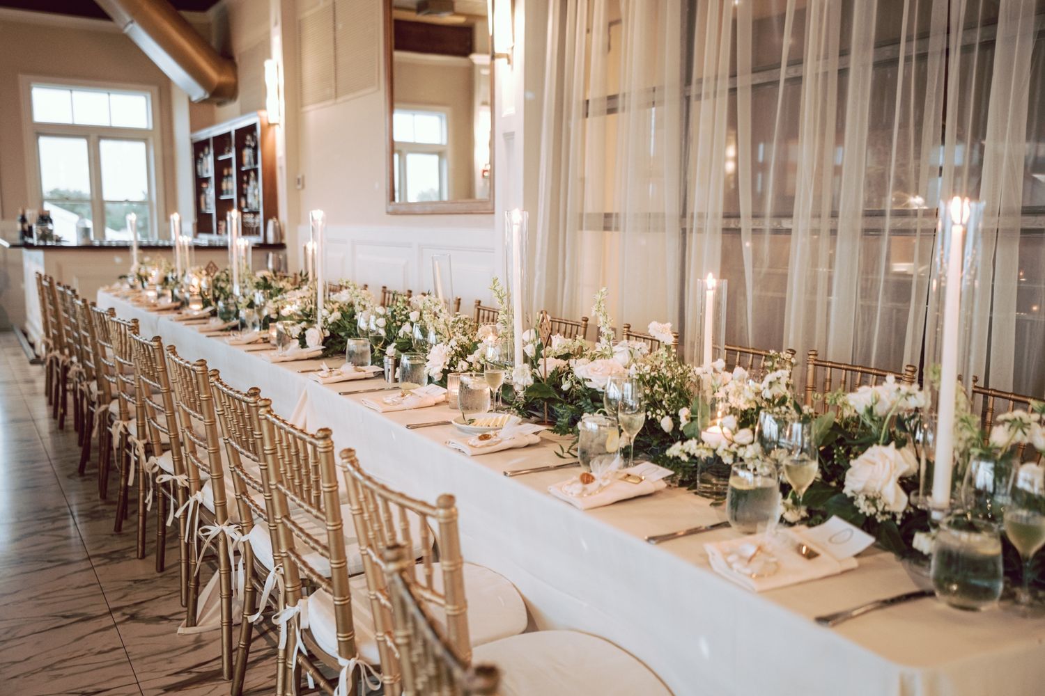 Elegant wedding reception long table with white linens, gold chiavari chairs, candles, and floral centerpieces in a bright venue.