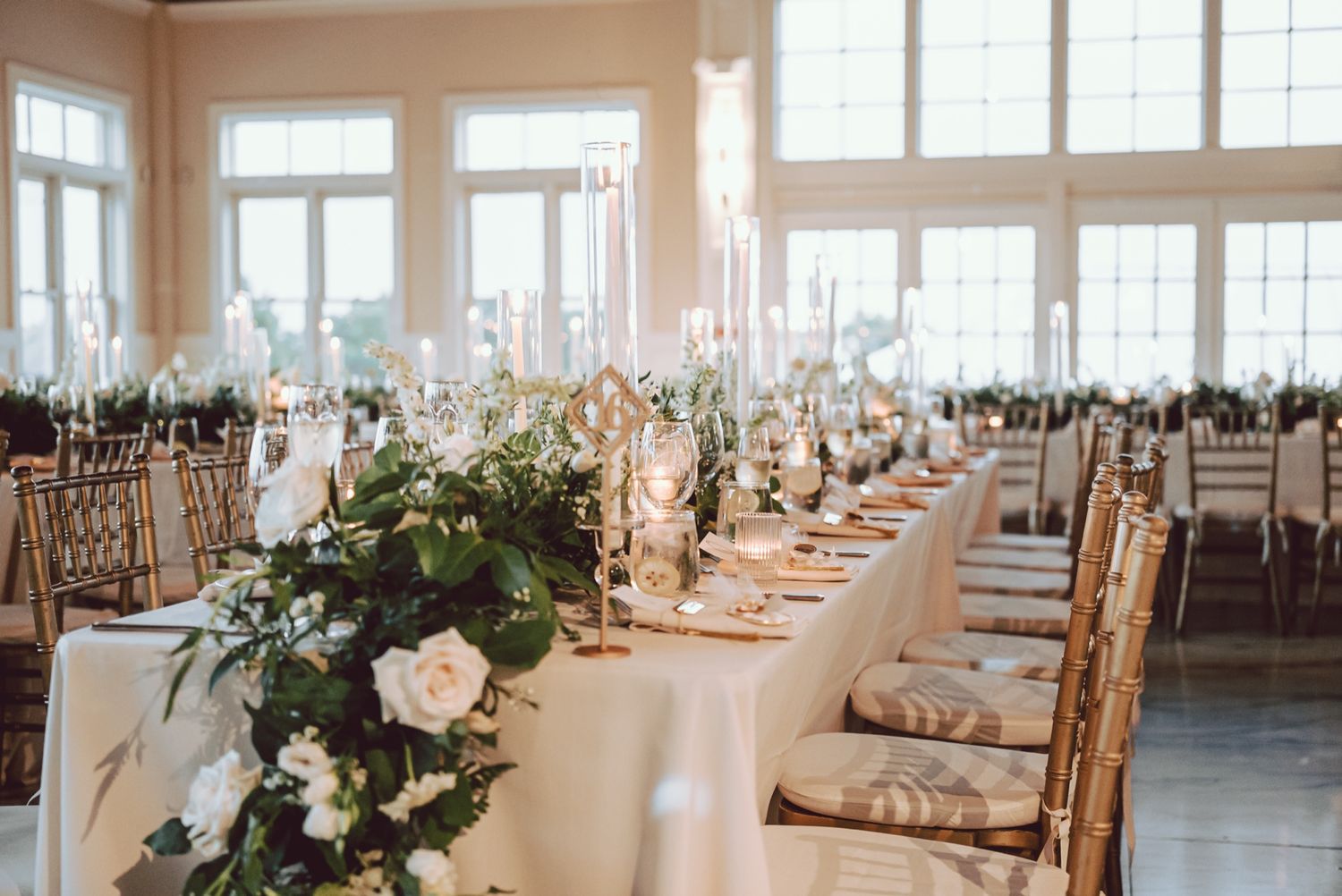Elegant wedding reception table with white floral runner, gold candelabras, and chiavari chairs in a bright venue.