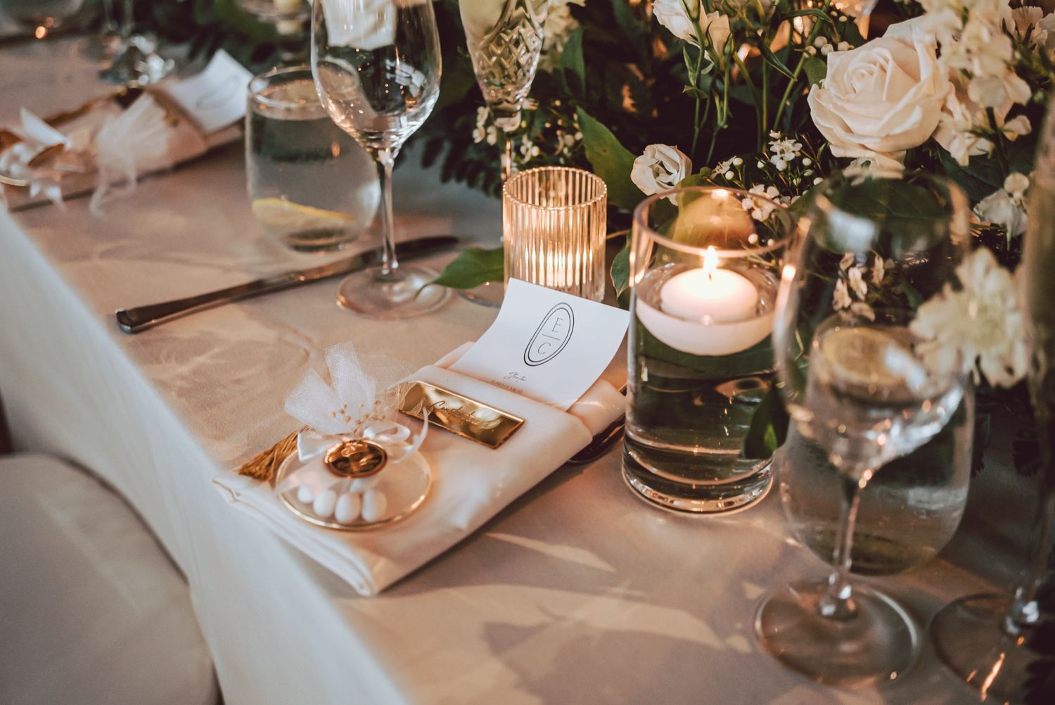 Elegant wedding table setting with white roses, floating candles, crystal glasses, and gold accents on a white linen tablecloth.