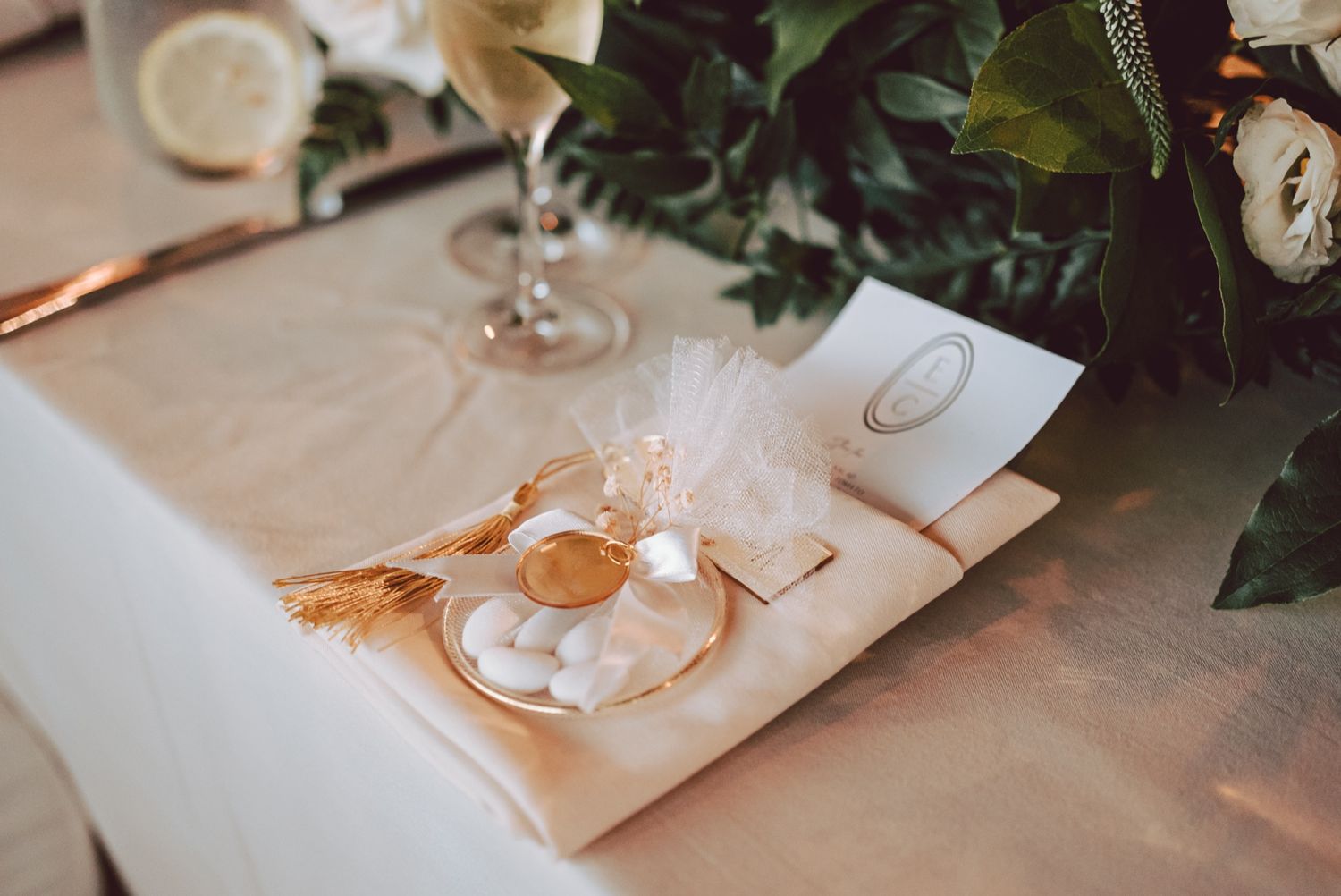 Elegant wedding table setting with white napkin, gold favor dish, sugared almonds, and floral greenery centerpiece.
