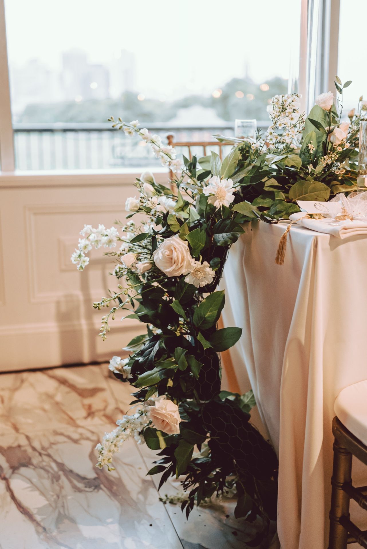 Elegant wedding table adorned with lush greenery and soft blush roses cascading down, set near a bright window.