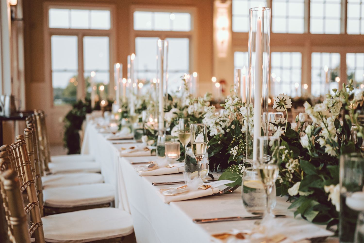 Elegant wedding reception table with white candles, lush greenery, and floral centerpieces in a bright venue.