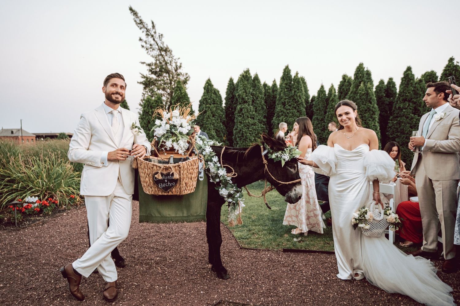 A bride and groom walk with a flower-adorned donkey at their outdoor wedding ceremony surrounded by guests.