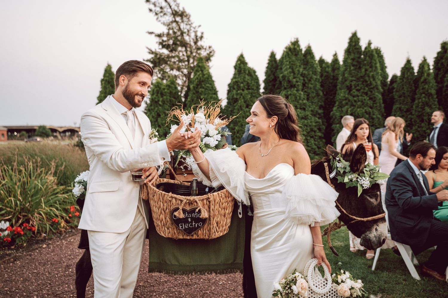 Bride and groom smiling while retrieving items from a decorated basket carried by a donkey at their wedding.