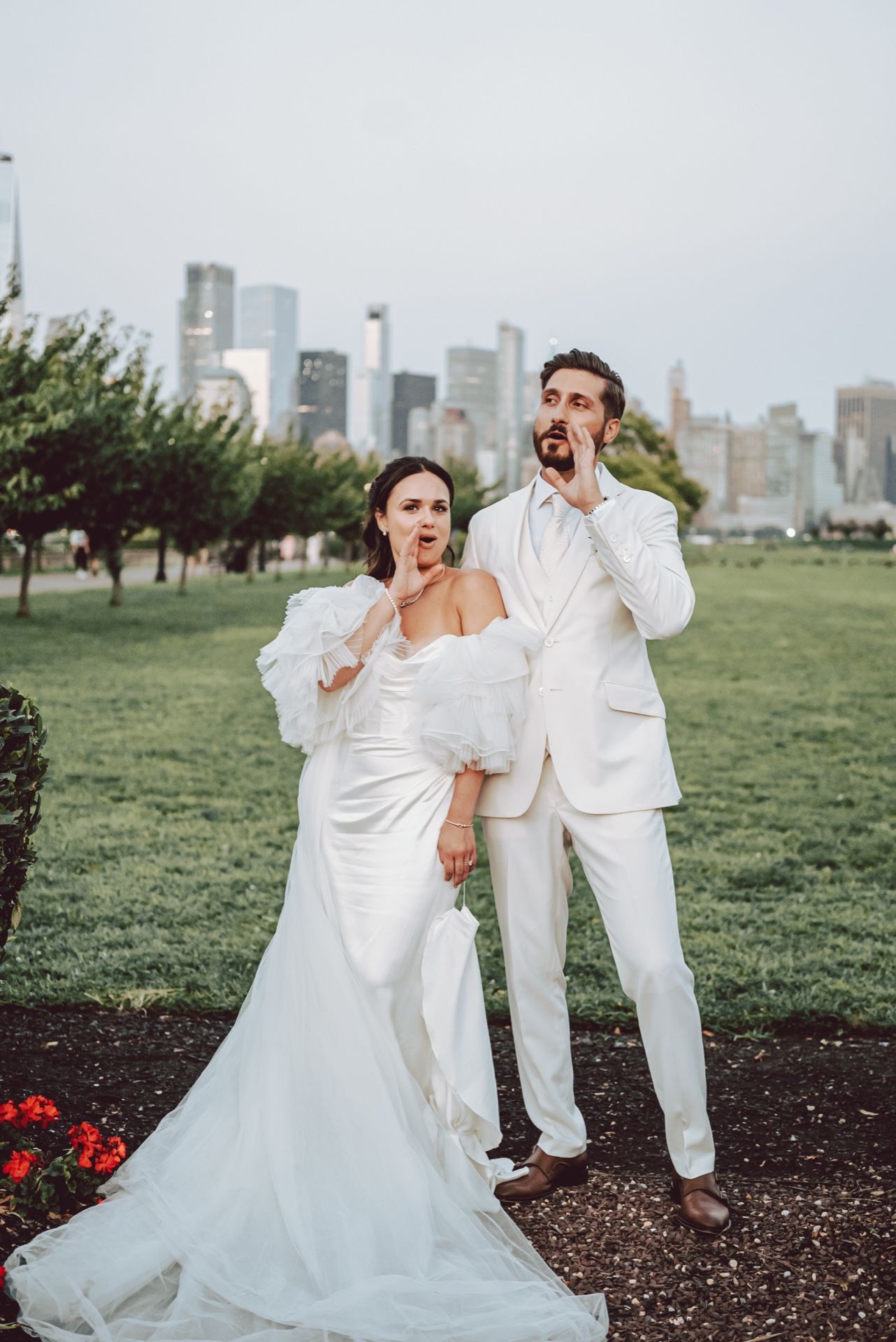 Bride and groom in white wedding attire strike a playful pose in a green park with a city skyline backdrop.