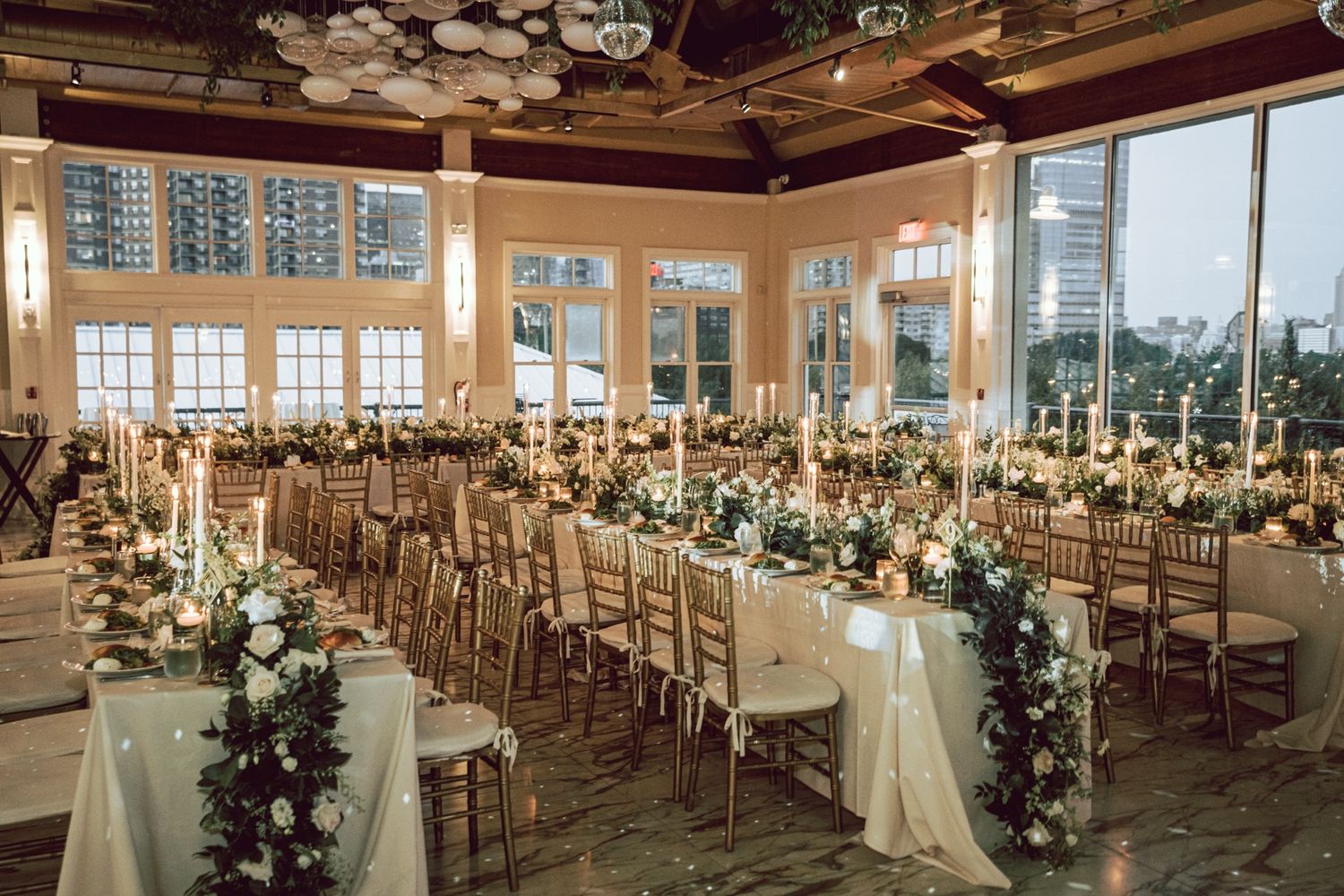 Elegant wedding reception hall with long tables, gold chairs, candles, and floral garlands in a windowed venue.