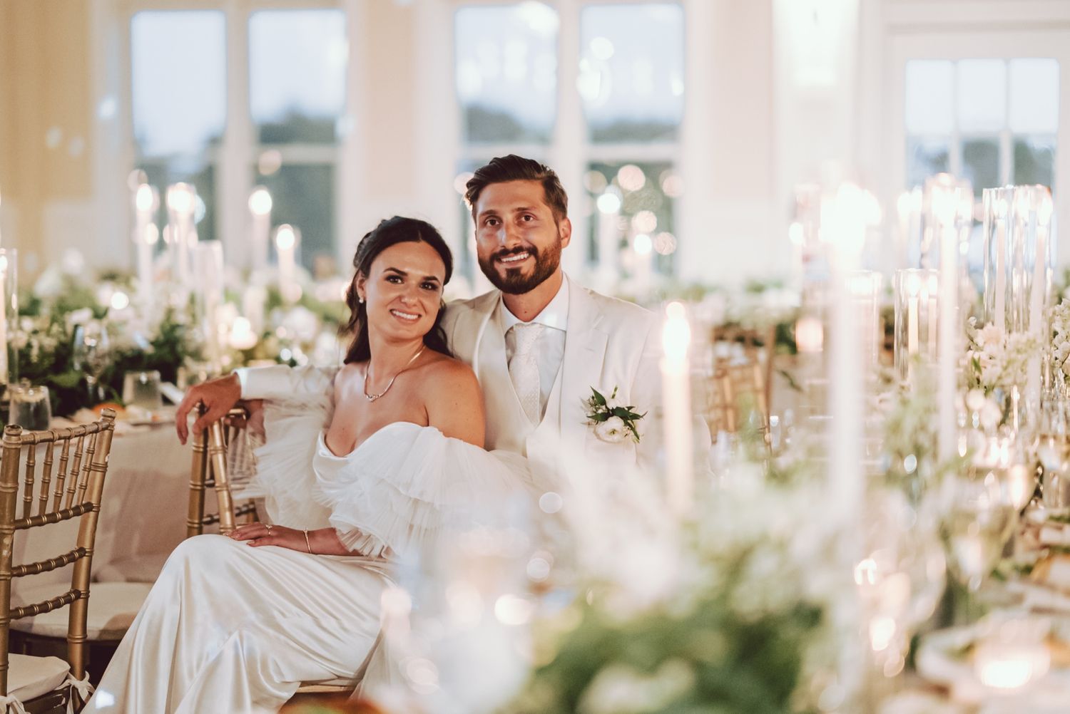 Bride and groom smile together at elegant candlelit wedding reception with floral centerpieces.