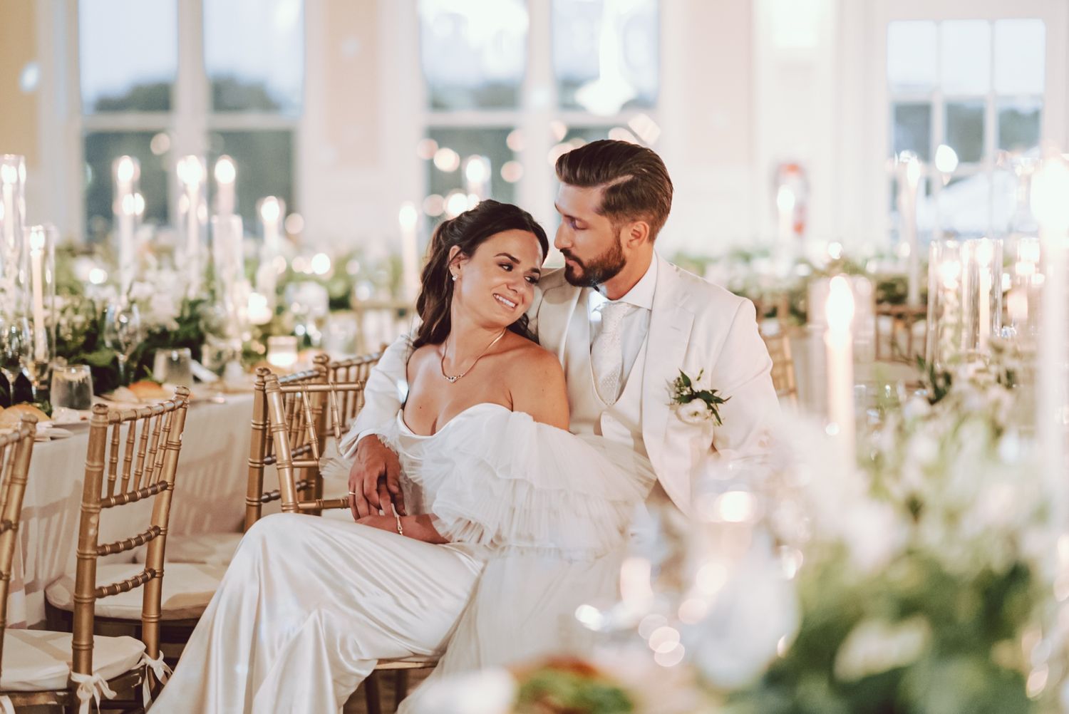 Bride and groom share an intimate moment seated together at elegant white floral wedding reception.