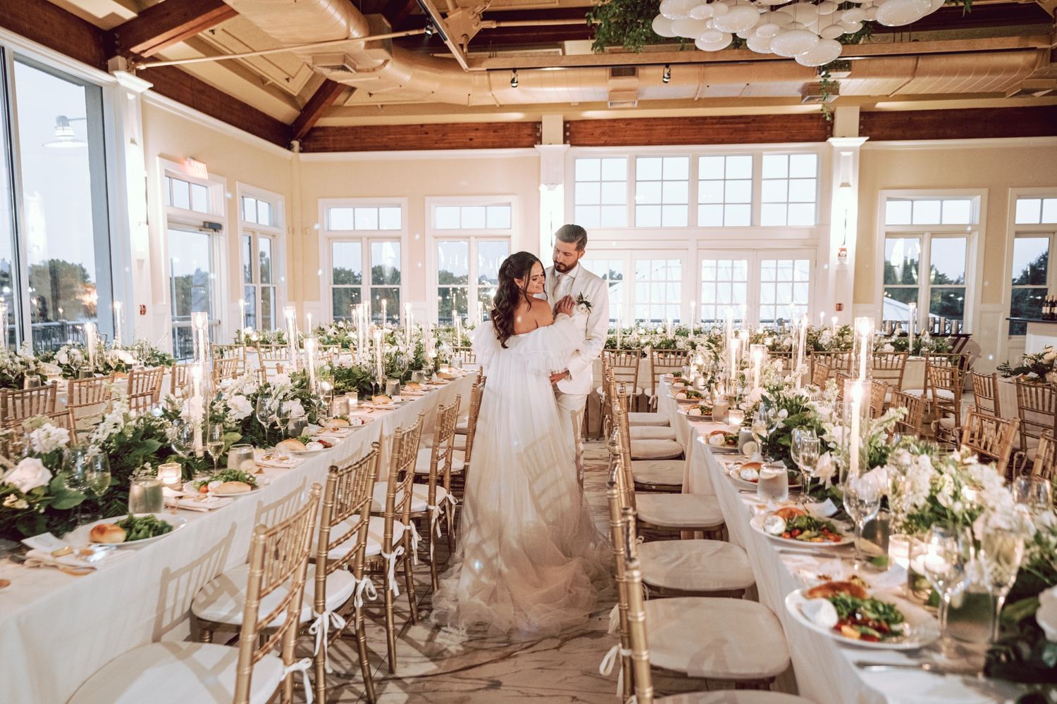 Couple stands together in stunning white wedding reception hall with long floral-adorned tables.