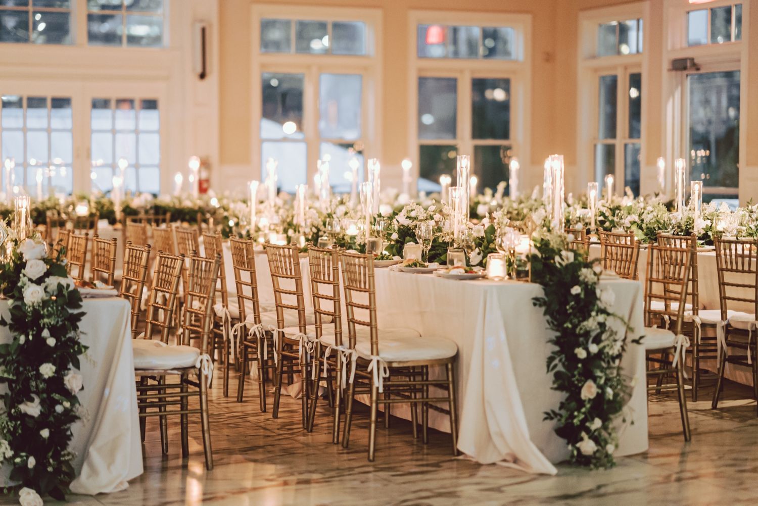 Elegant wedding reception hall with long tables, gold chiavari chairs, candles, and floral arrangements.