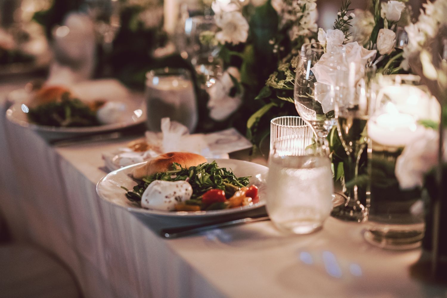 Elegant wedding reception table with floral centerpieces, white roses, candles, and a plated salad dish.