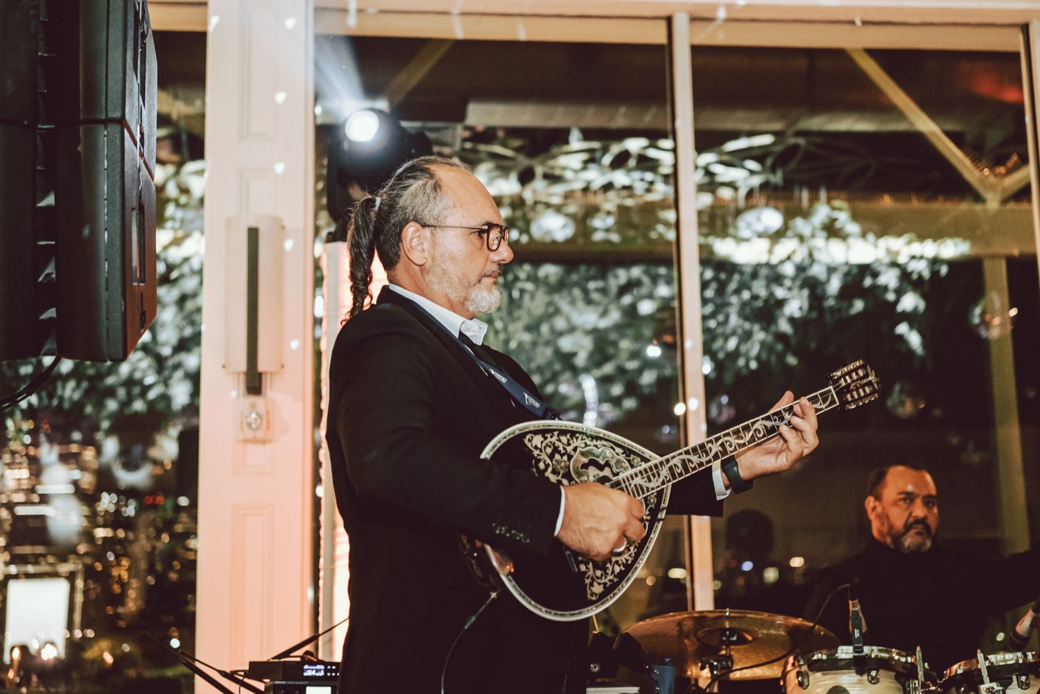 Older man with glasses playing a mandolin in a black suit at an elegant indoor evening event with a drummer behind him.