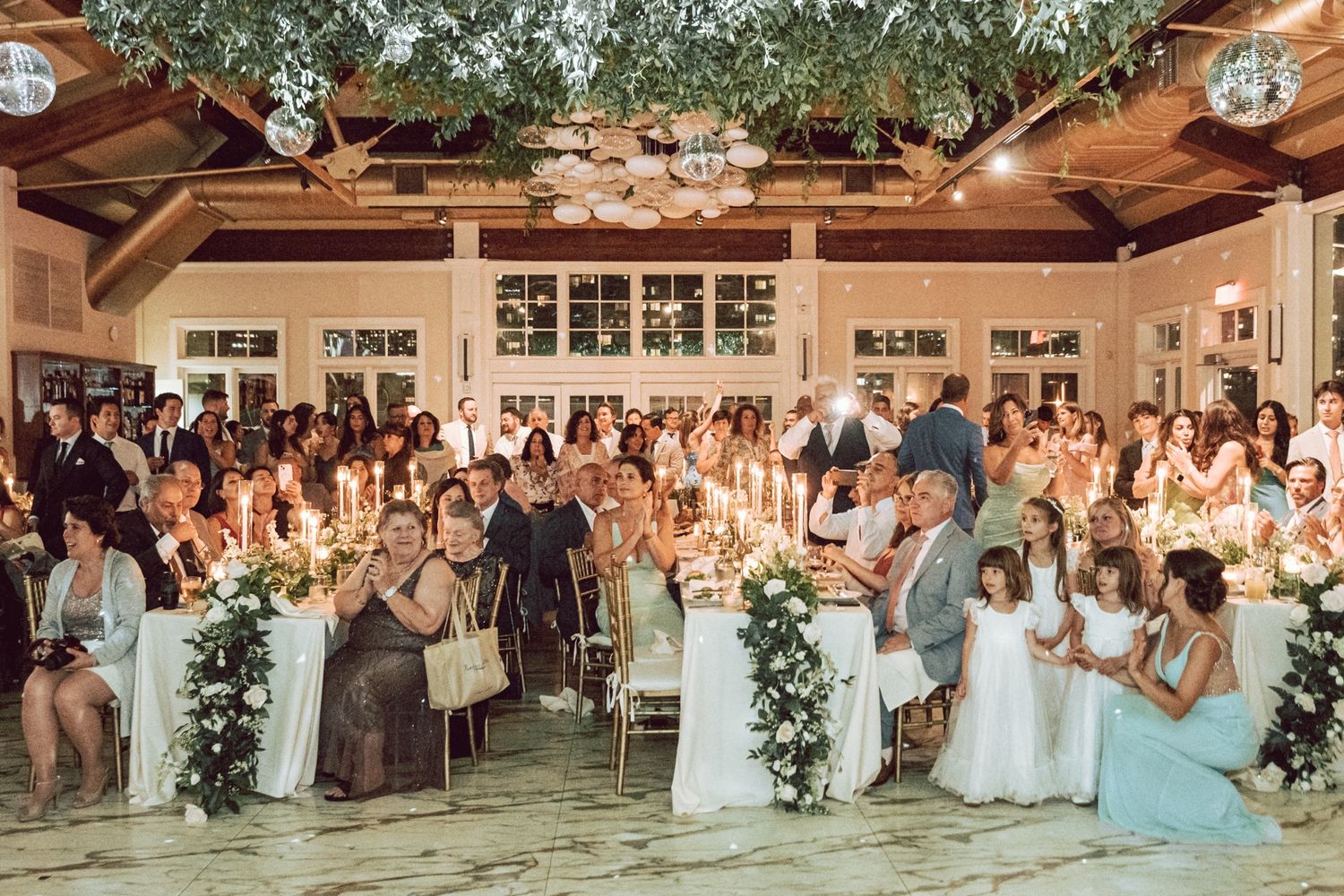 Elegant wedding reception with guests seated at candlelit tables adorned with white floral arrangements in a warmly lit venue.