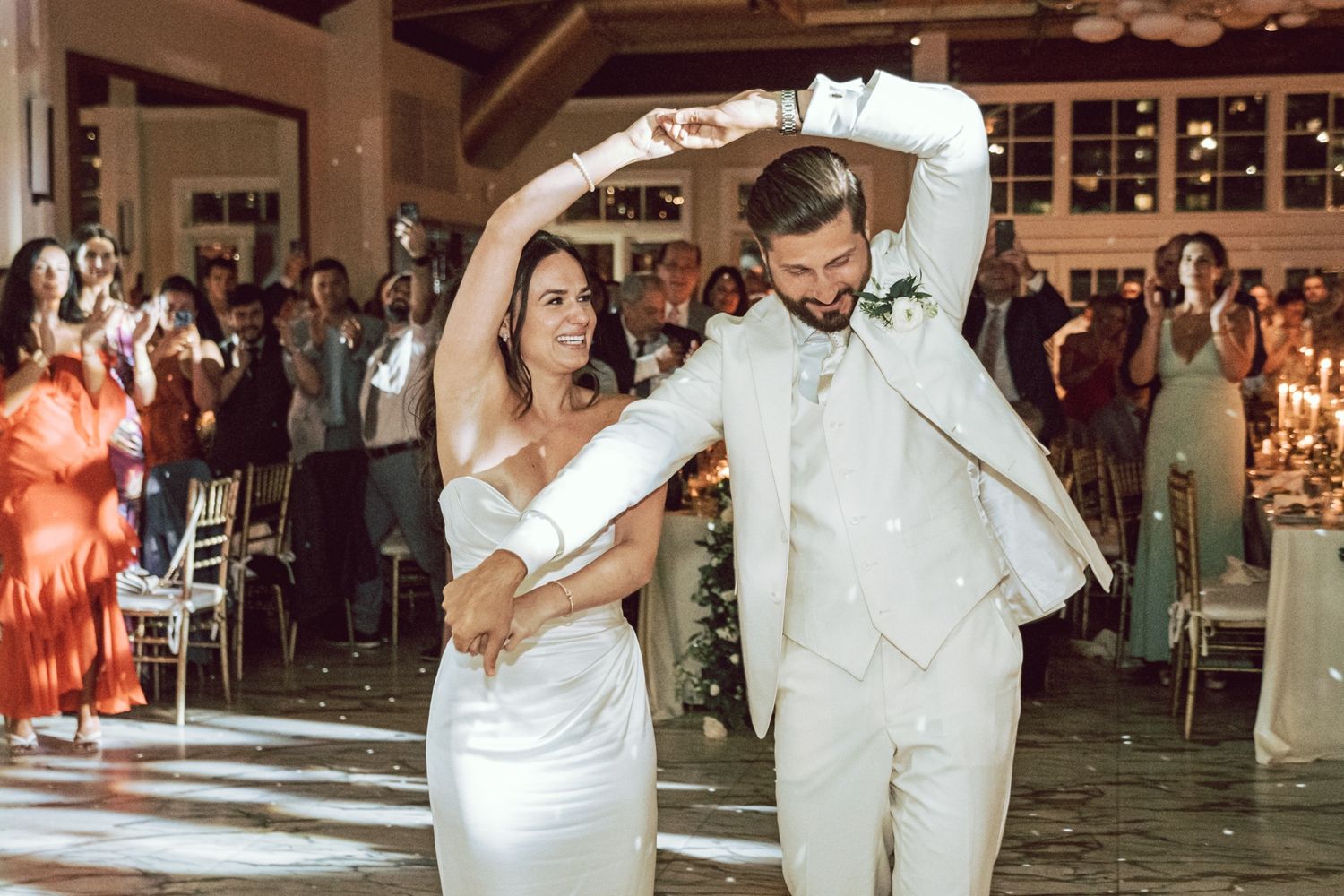 Bride and groom share first dance at wedding reception, guests applauding in background.