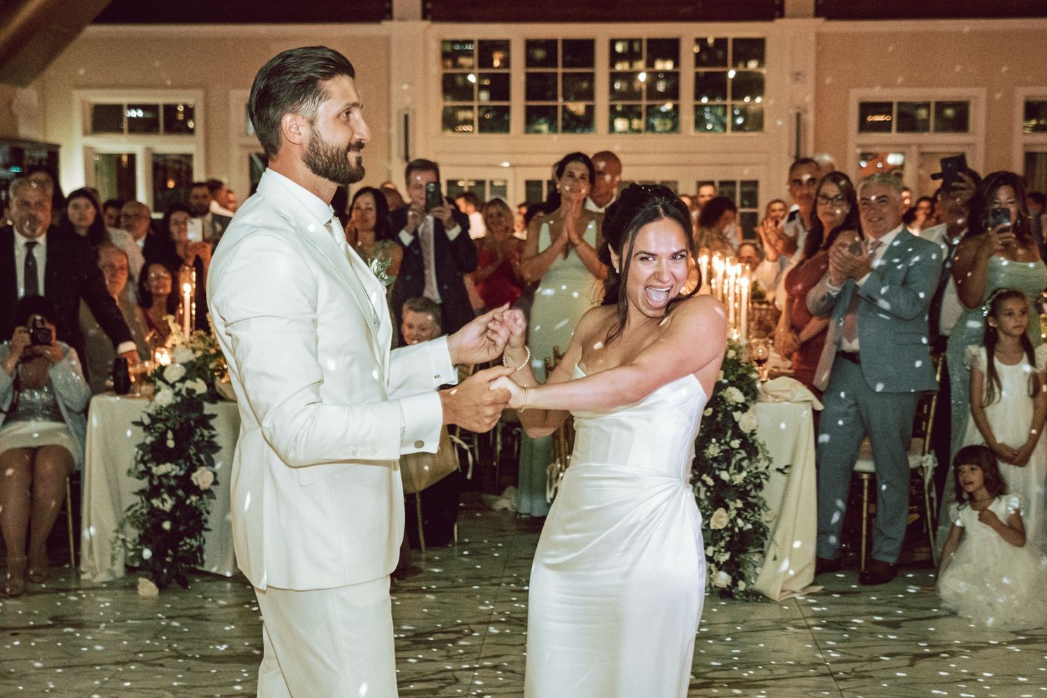 Bride and groom share first dance at elegant wedding reception with candlelit backdrop and cheering guests.