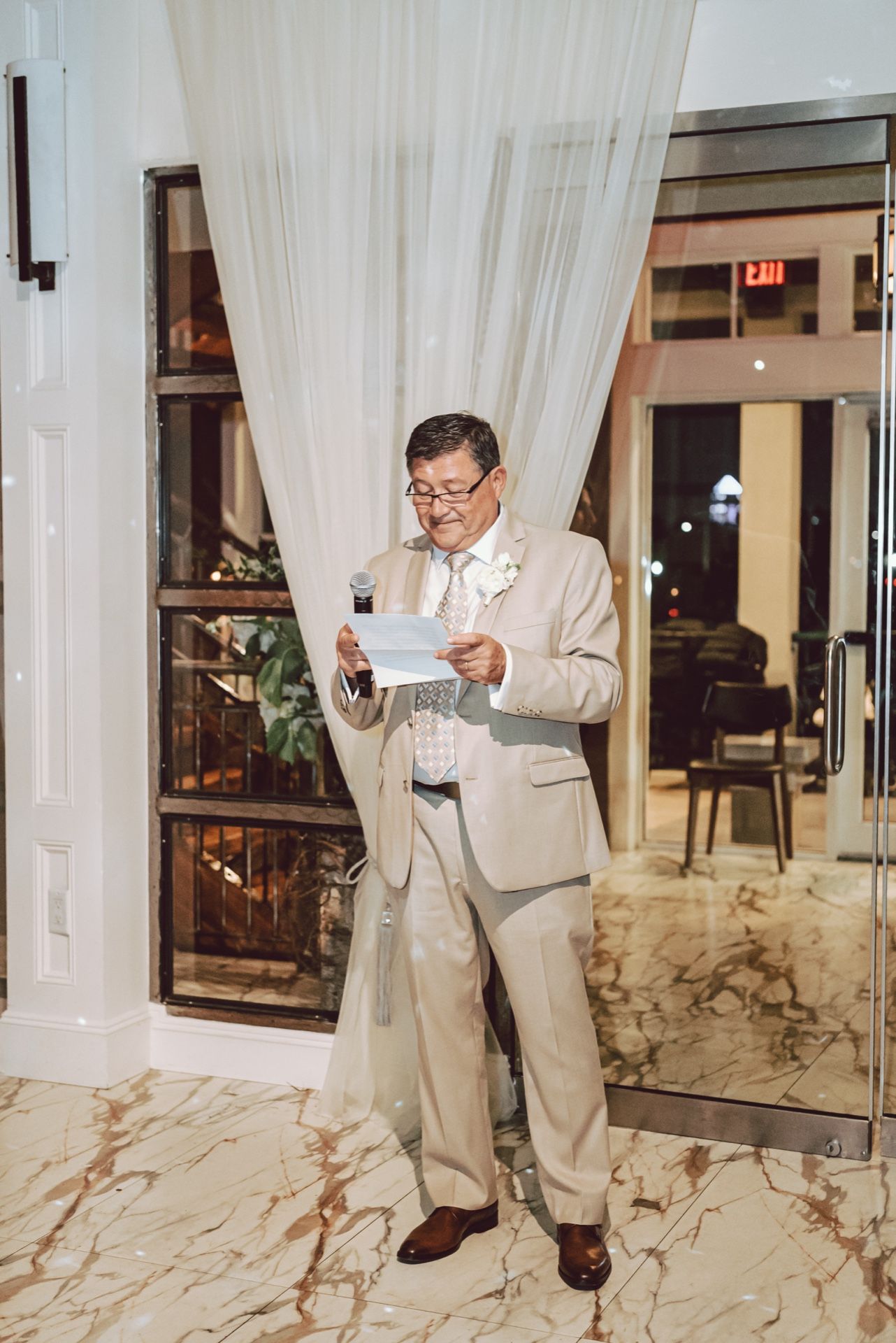 Man in beige suit reads speech at wedding reception, holding microphone and paper near draped curtains on marble floor.