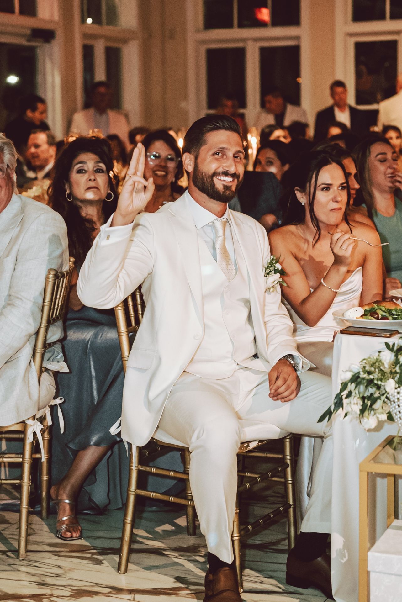 Smiling groom in white suit raises hand at elegant wedding reception surrounded by seated guests.