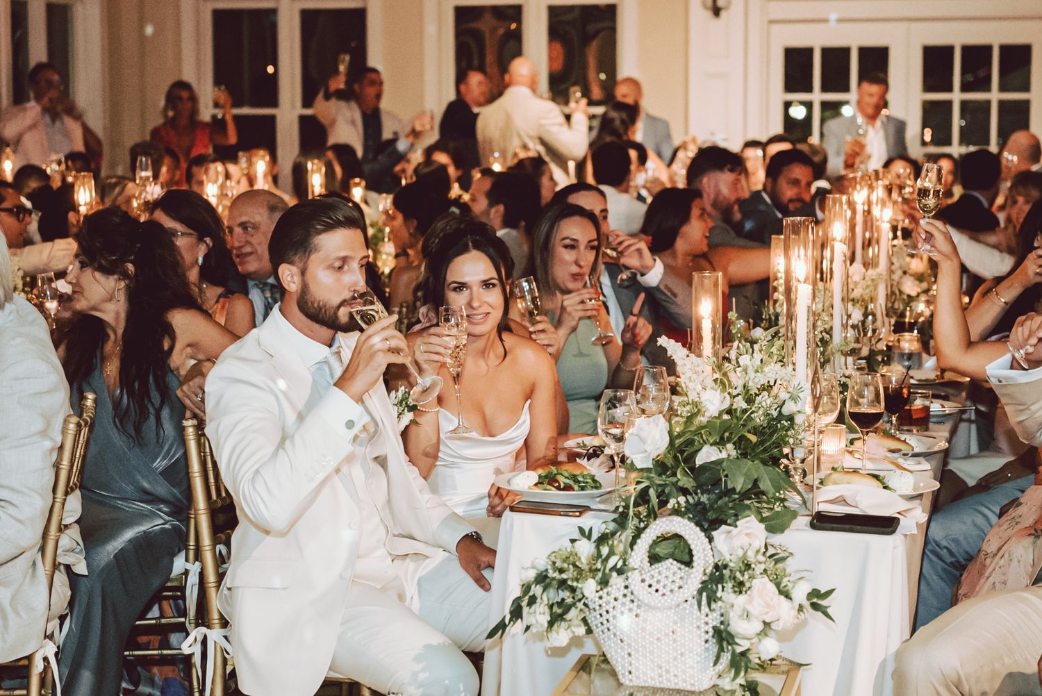 Elegant wedding reception with guests seated at candlelit tables adorned with greenery, bride and groom sharing a romantic moment.