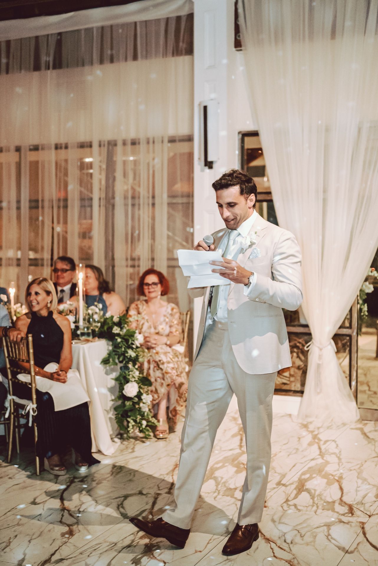 Groom in light gray suit reads vows at elegant wedding reception with floral decor and guests seated in background.