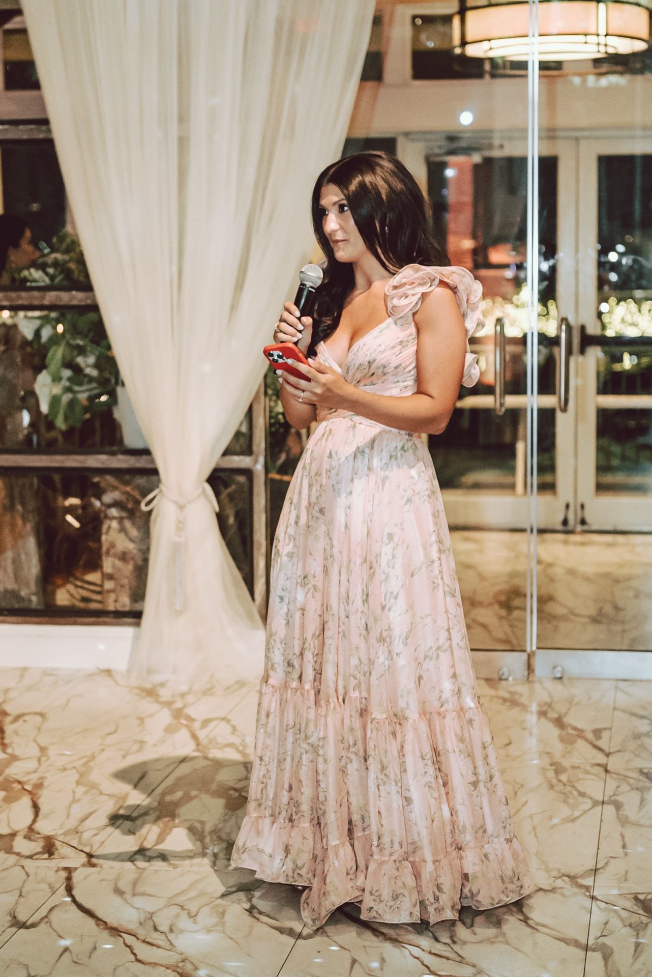 Woman in floral pink gown speaks into microphone at elegant wedding reception venue.