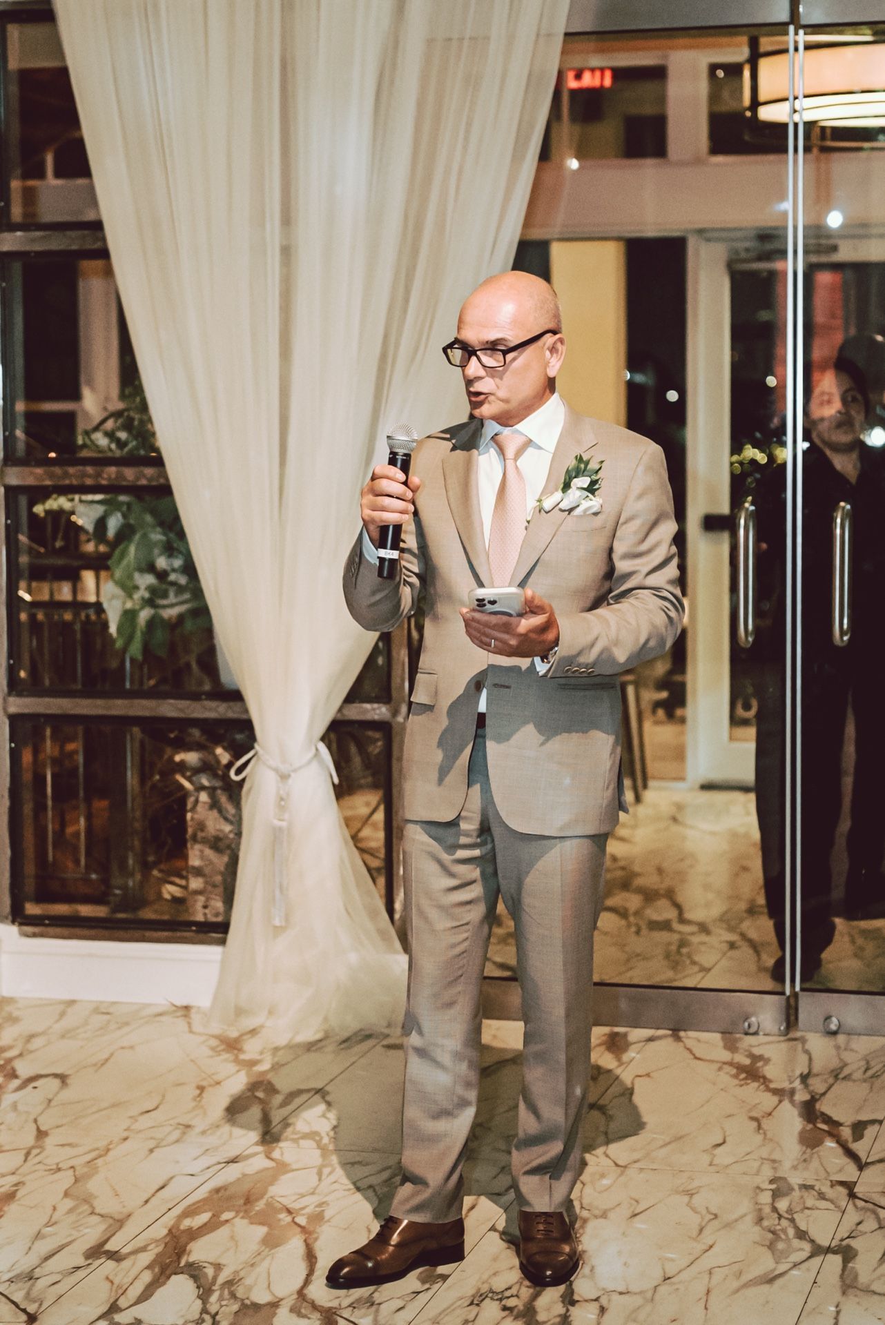 Bald man in tan suit delivers speech at microphone during wedding reception.
