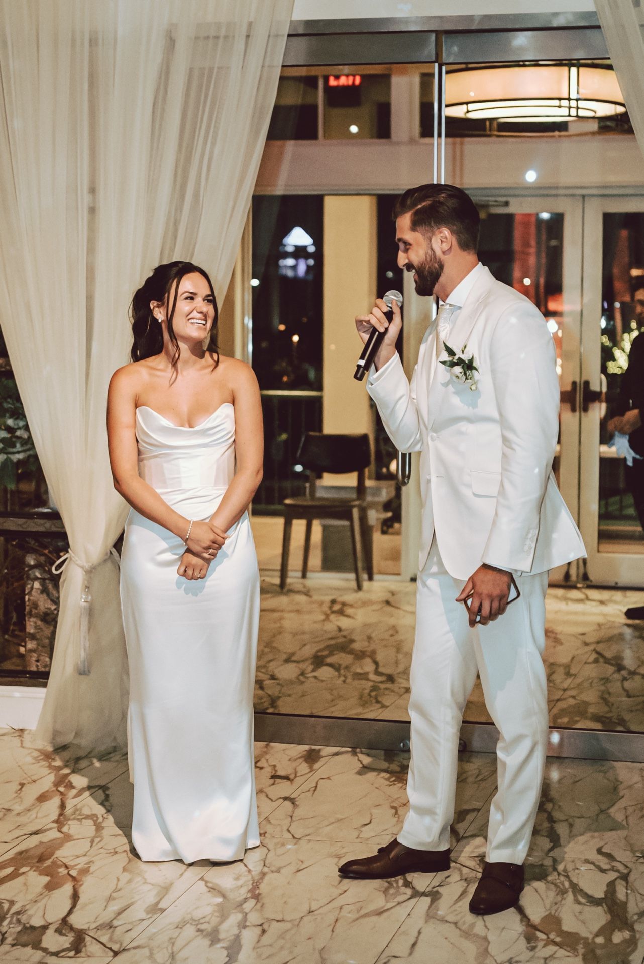 Groom in white suit speaks into microphone while bride in satin gown smiles during wedding reception speech.