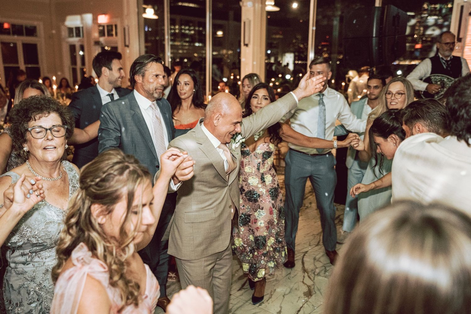 Guests dancing joyfully at an elegant evening wedding reception, with the groom in a tan suit leading the celebration.