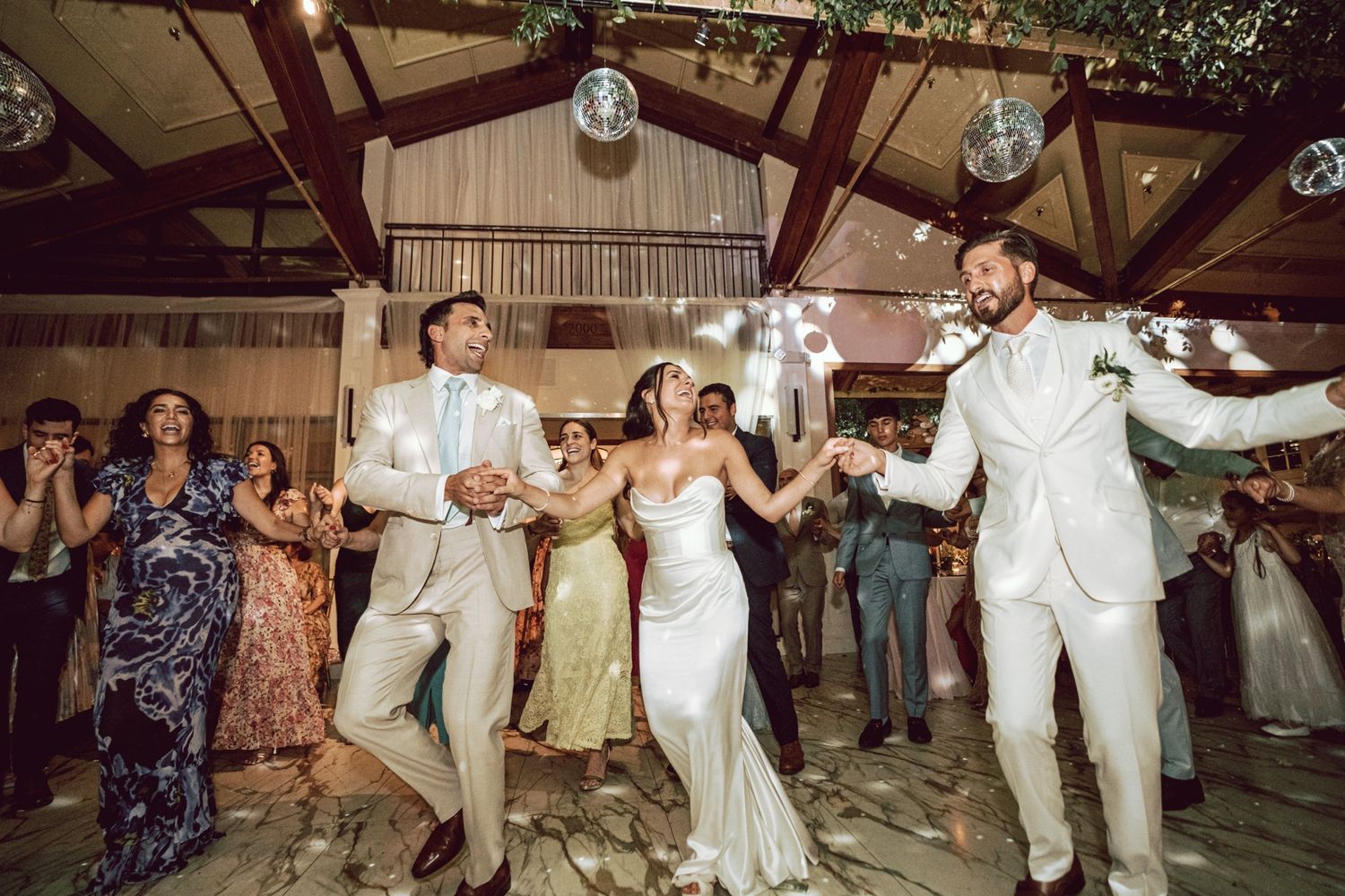 Bride and groom dancing joyfully with guests at elegant indoor wedding reception with disco ball lighting.