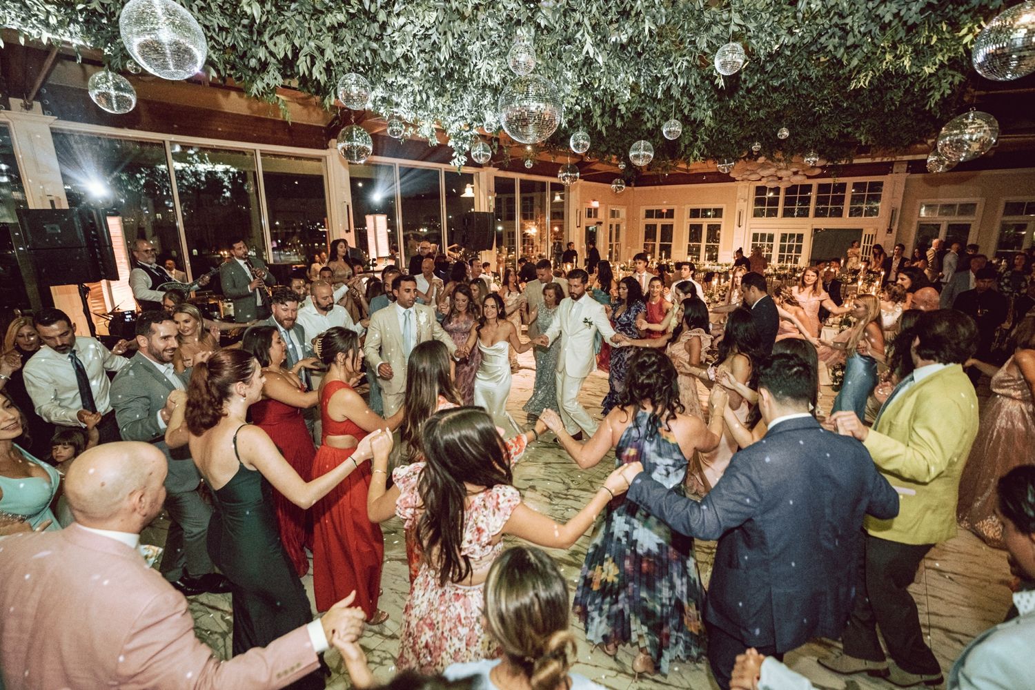 Guests dancing joyfully at an elegant indoor wedding reception with disco balls and lush greenery decor.