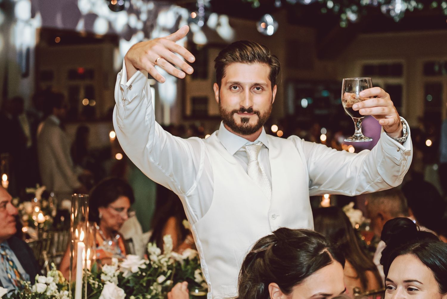 Groomsman in white shirt raising a glass for a toast at a romantic candlelit wedding reception.