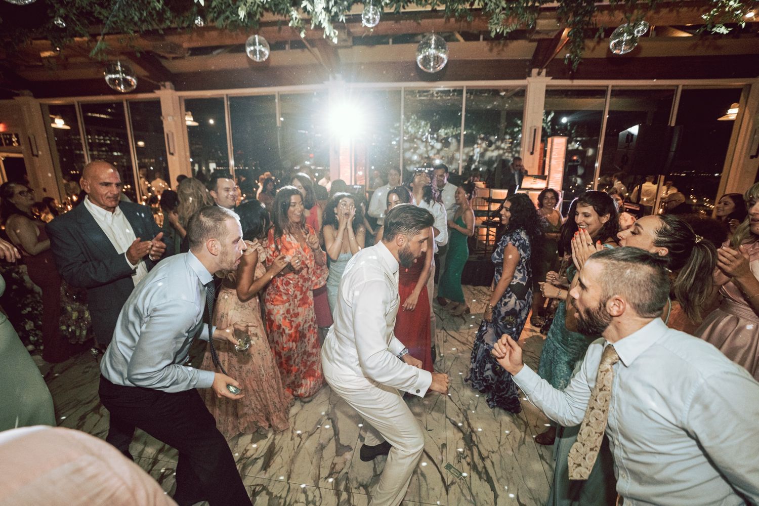 Guests celebrate on the dance floor at a lively indoor wedding reception with festive lighting.