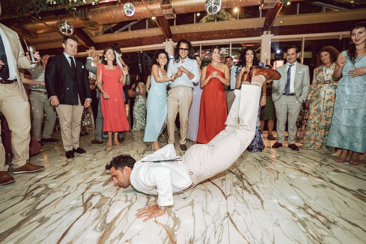 Groom in white suit breakdancing on the dance floor surrounded by cheering wedding guests.