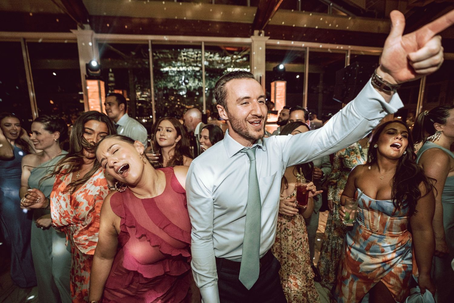 Guests dancing and celebrating joyfully at a lively wedding reception, man giving thumbs up in foreground.