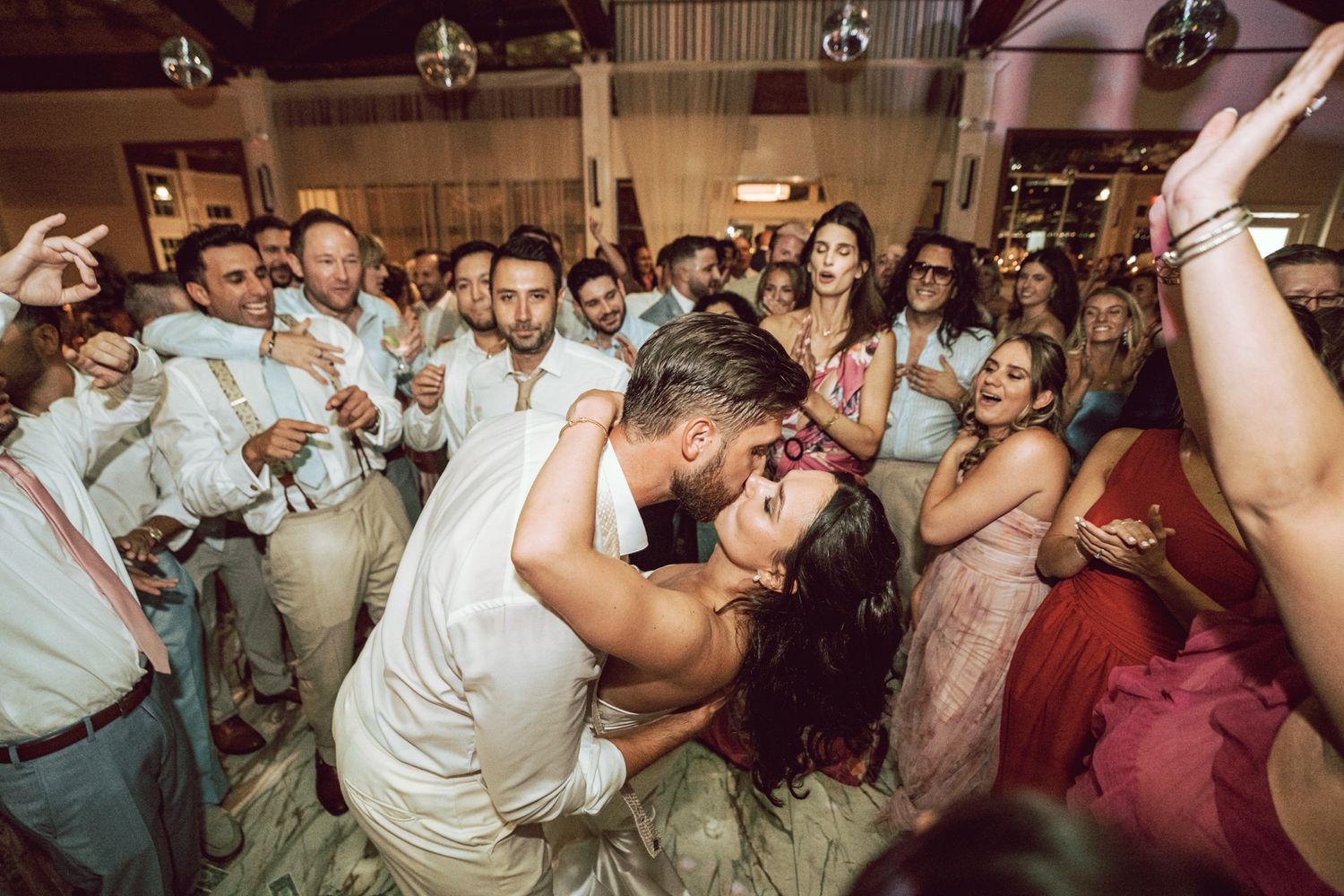 Bride and groom share a romantic dip kiss on the dance floor surrounded by cheering wedding guests.