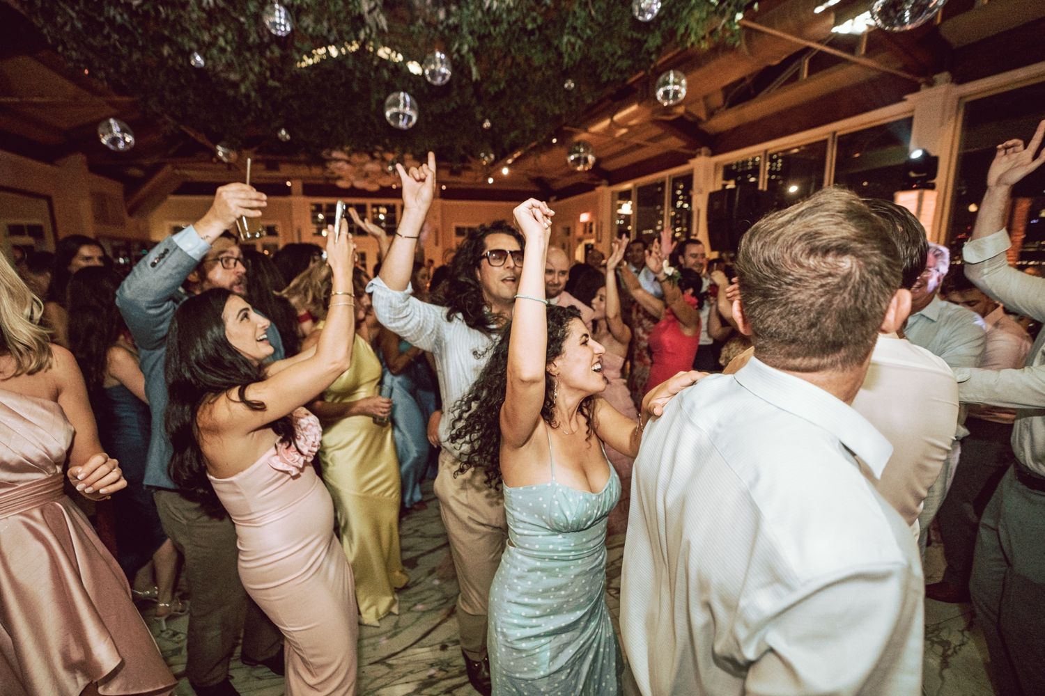 Wedding guests dance joyfully under disco balls and greenery at an elegant indoor reception.