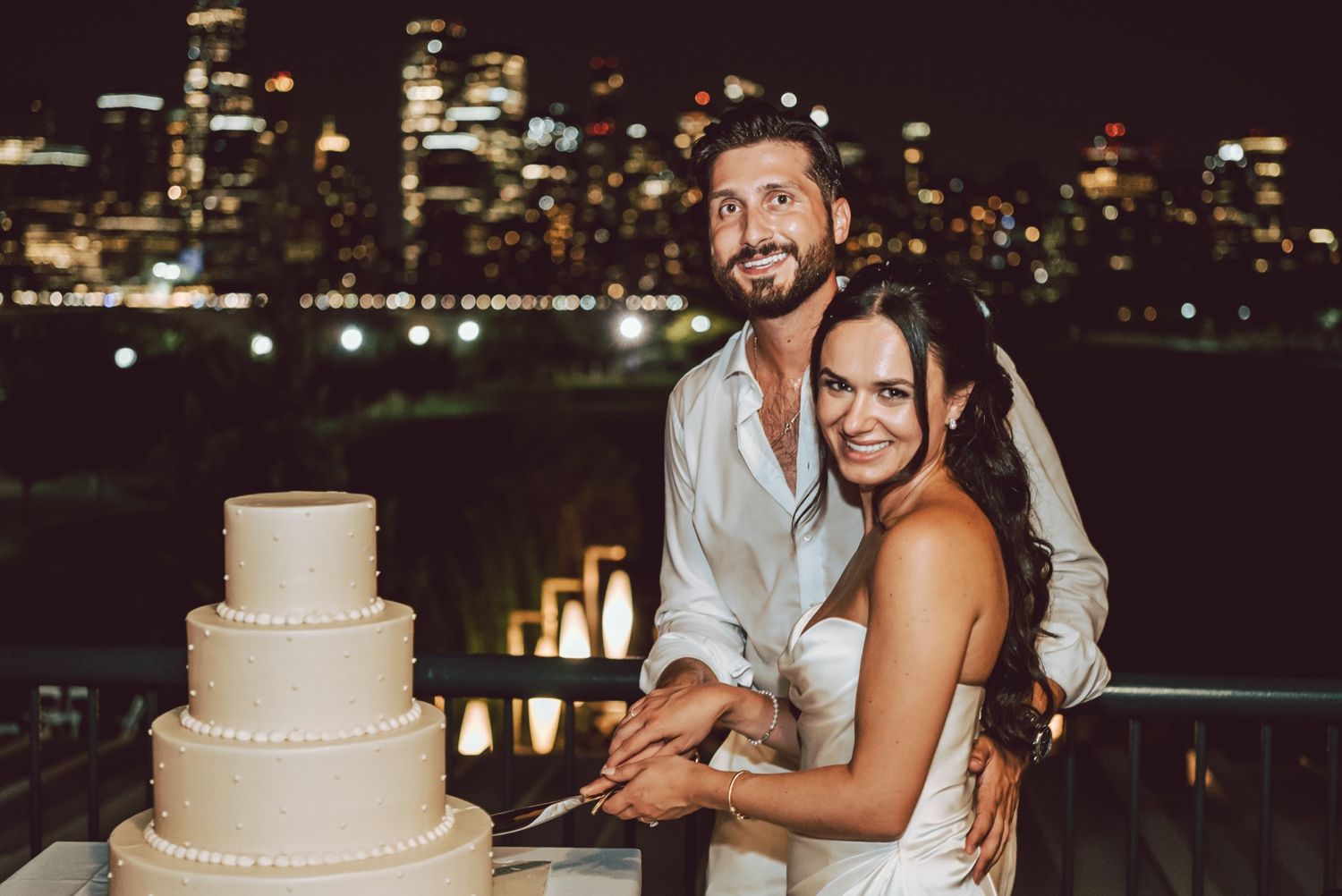 Bride and groom cutting wedding cake with city skyline at night.