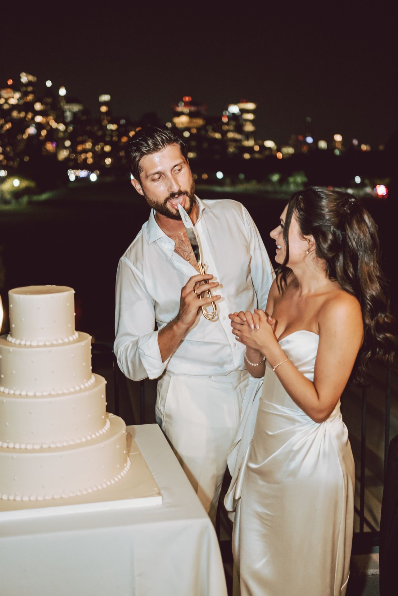 Bride and groom cutting wedding cake at night with city lights backdrop, dressed in elegant white attire.
