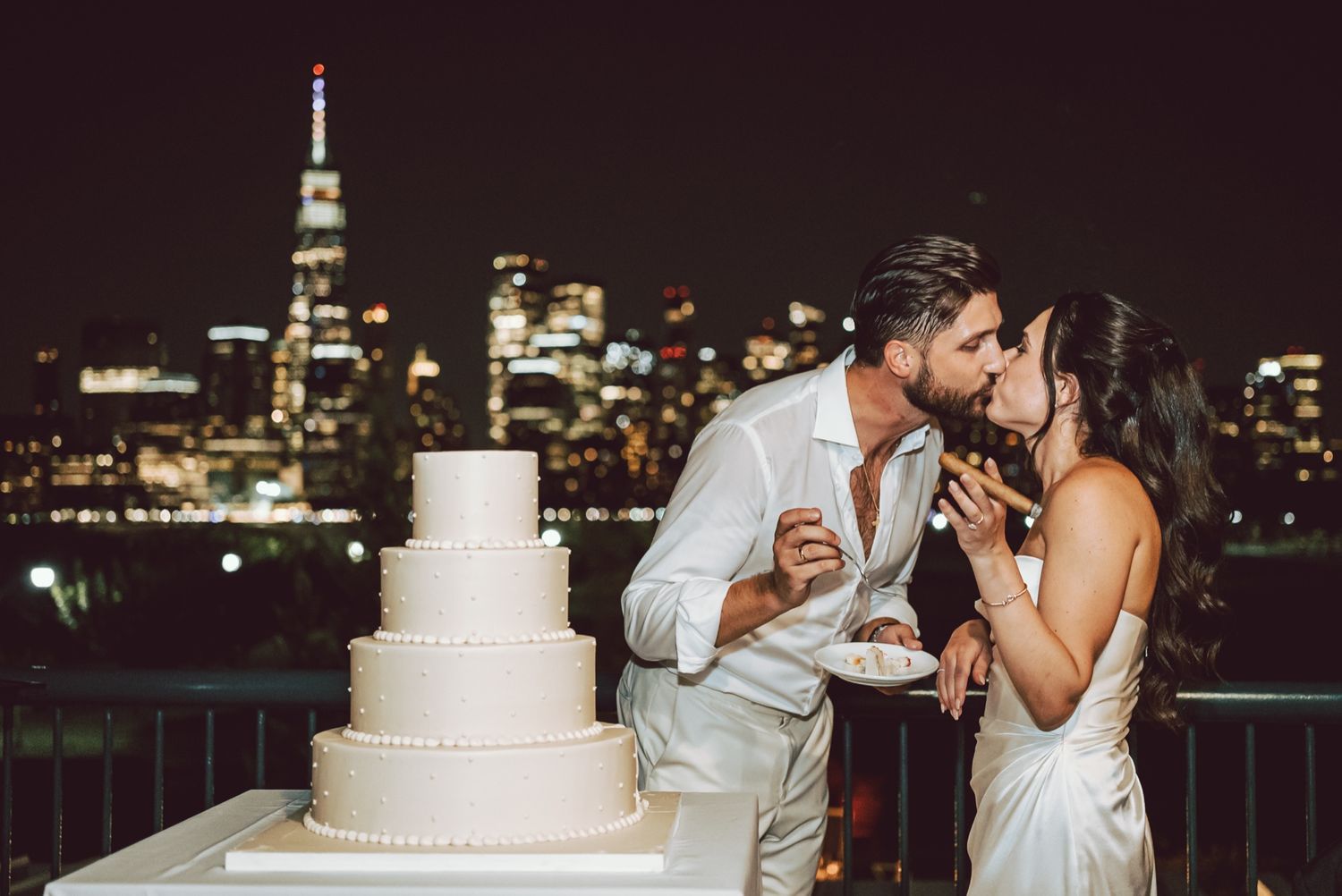 Newlyweds share a kiss while cutting their white wedding cake with NYC skyline at night.