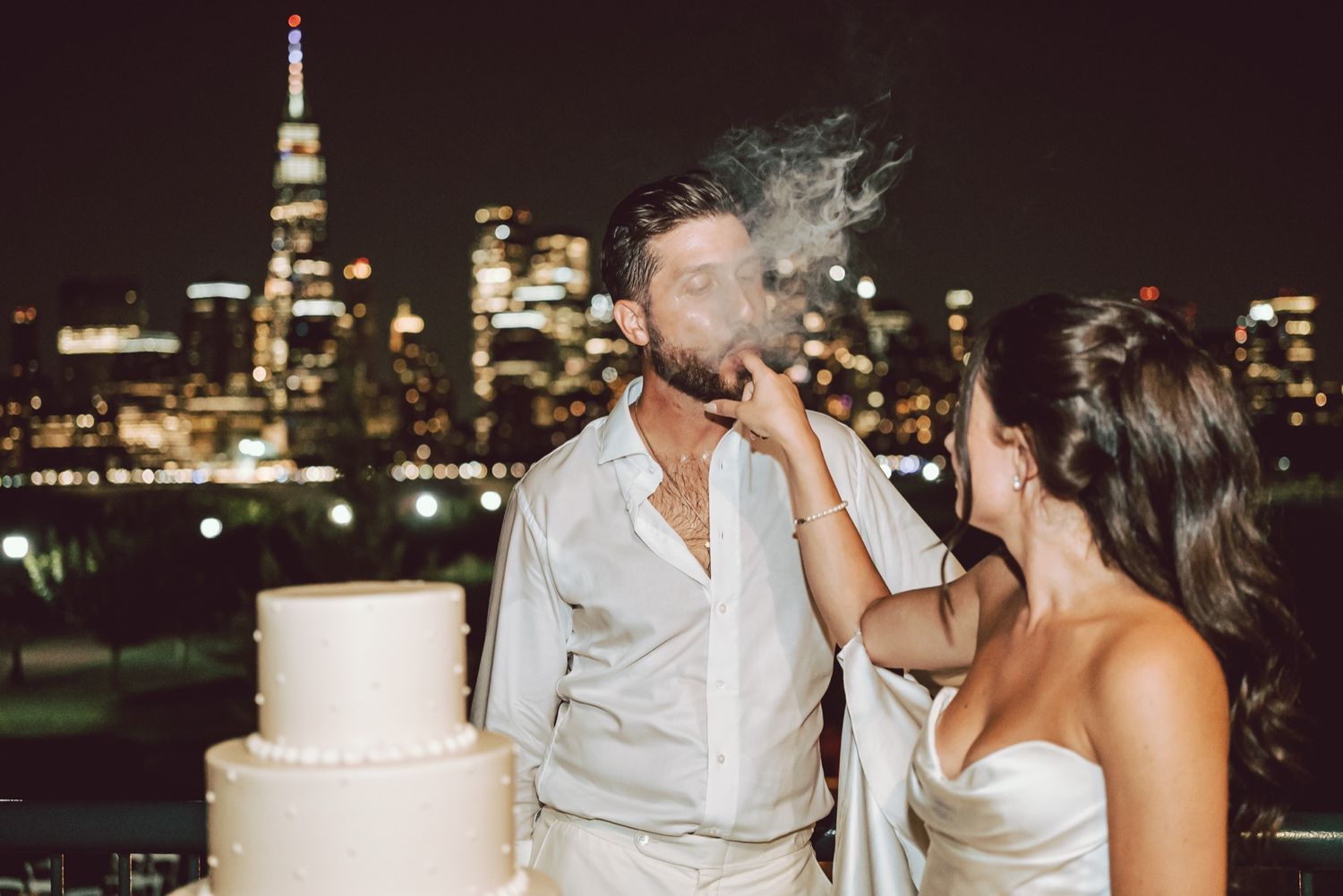 Couple cutting wedding cake on rooftop with NYC skyline at night, groom smoking cigar in white suit.