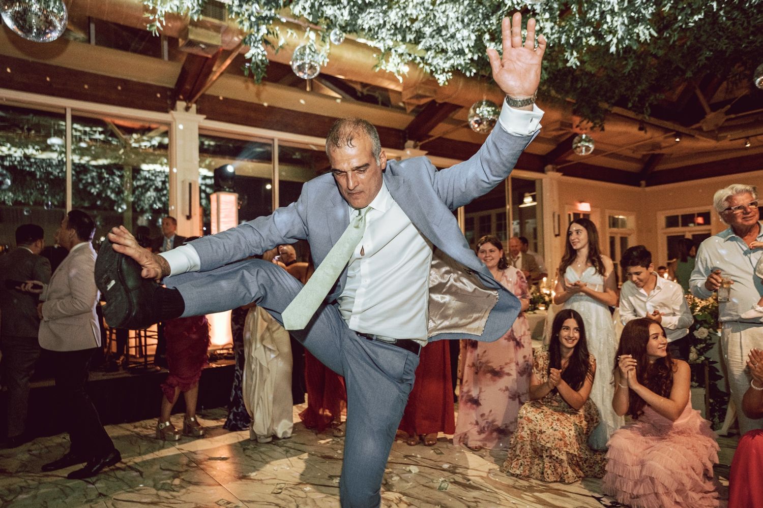 Man in blue suit dancing energetically at a wedding reception with guests watching and celebrating in the background.