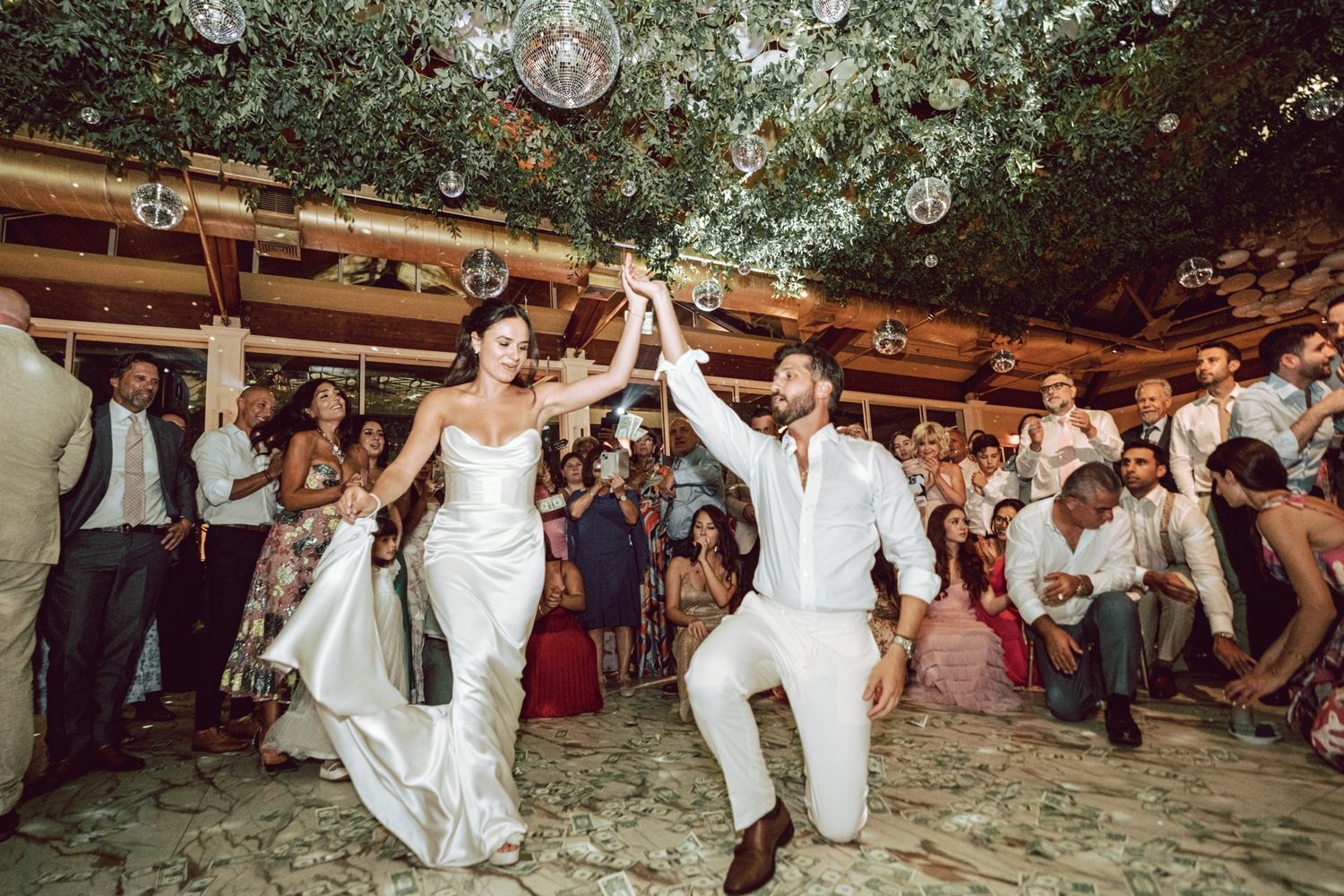 Bride and groom share first dance at elegant wedding reception with lush greenery ceiling and guests celebrating.