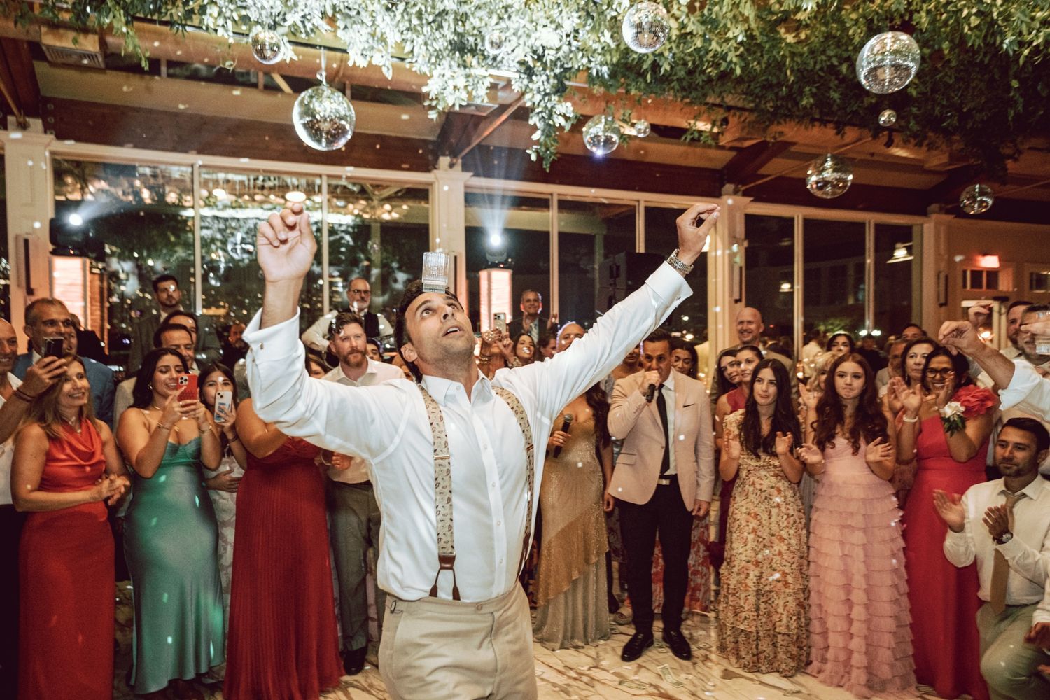 Groom triumphantly raises arms at wedding reception while guests celebrate on the dance floor.