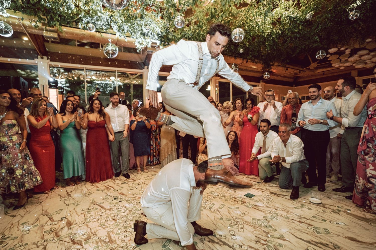 Groom leaping over a crouching guest on the dance floor at a lively outdoor wedding reception.