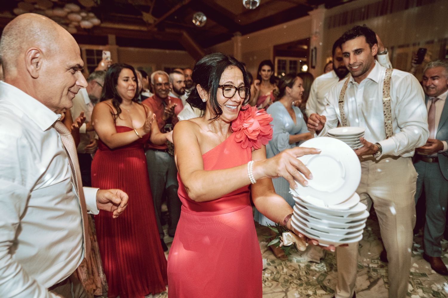 Woman in red dress smashing plates at a lively wedding reception celebration with guests cheering around her.
