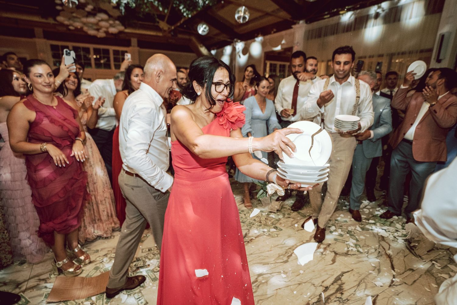 Woman in red dress smashing plates at lively wedding celebration with guests watching.