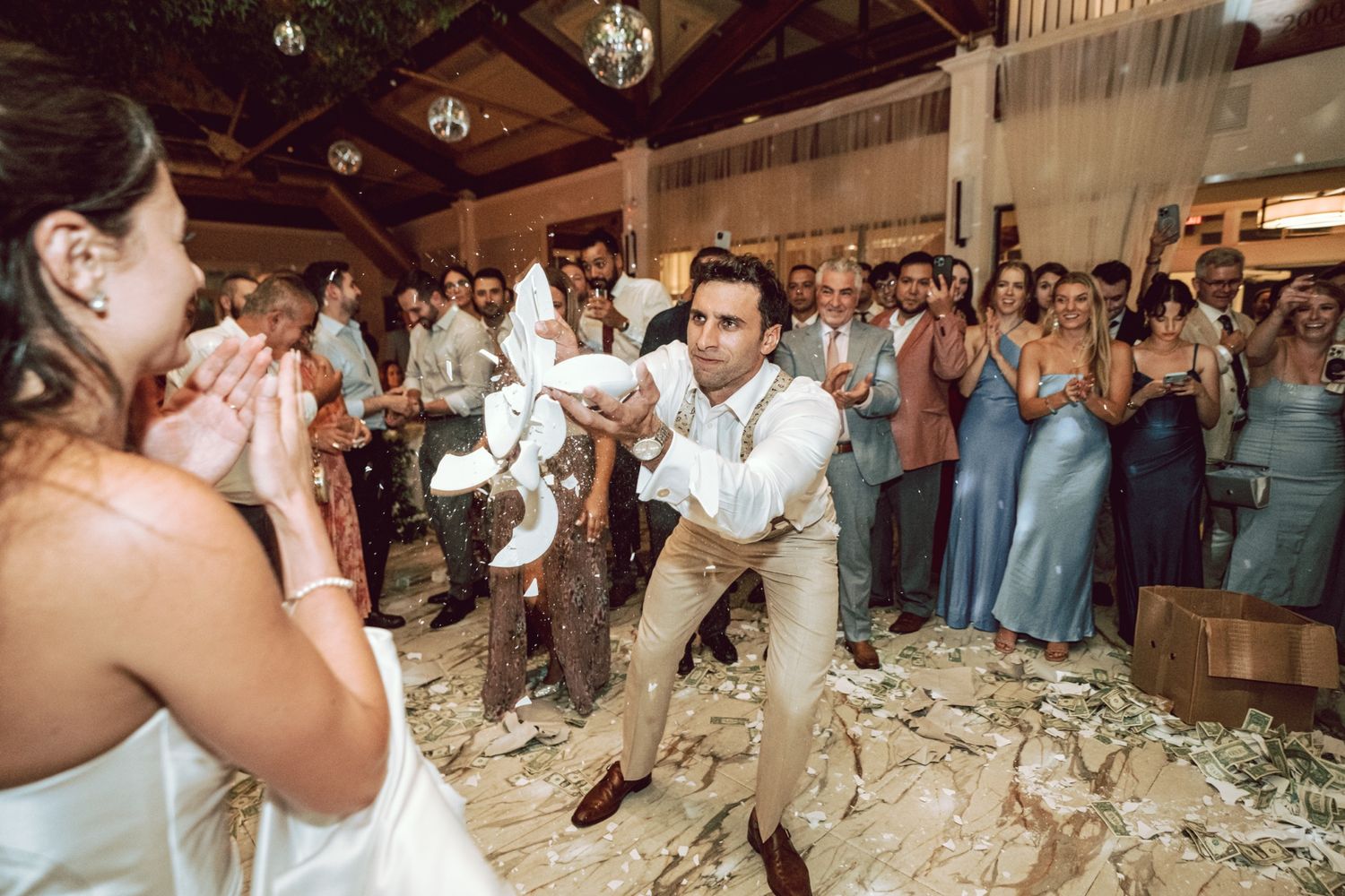 Groom tossing money at a lively wedding reception while guests clap and celebrate on the dance floor.
