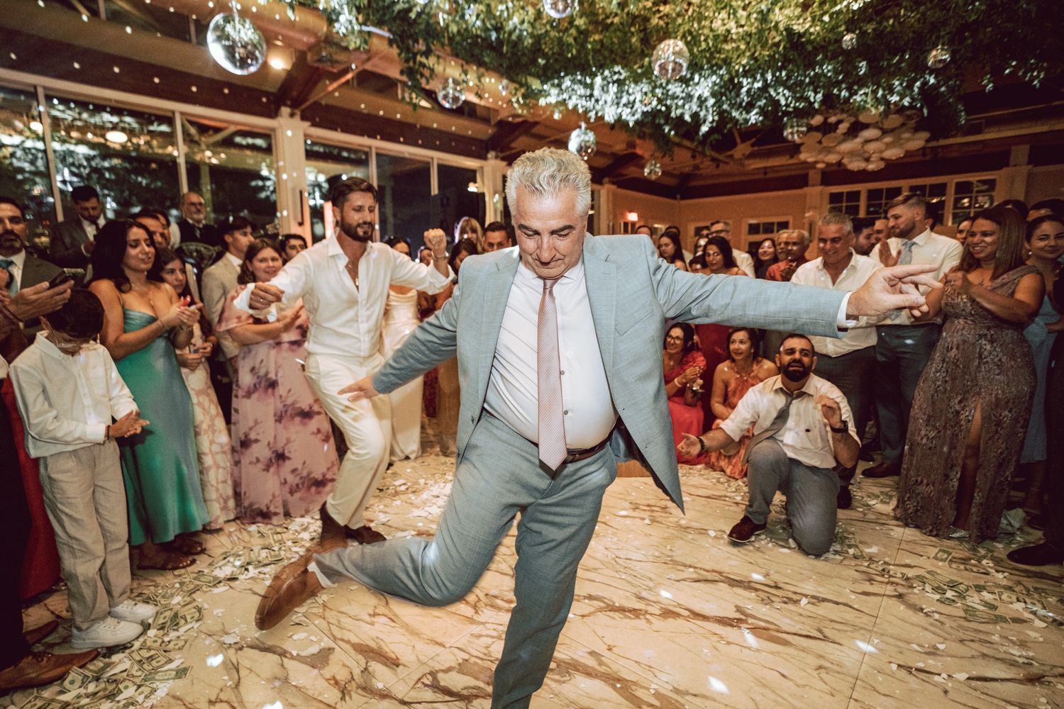 Man in light blue suit energetically dancing at a lively wedding reception with guests cheering around him.