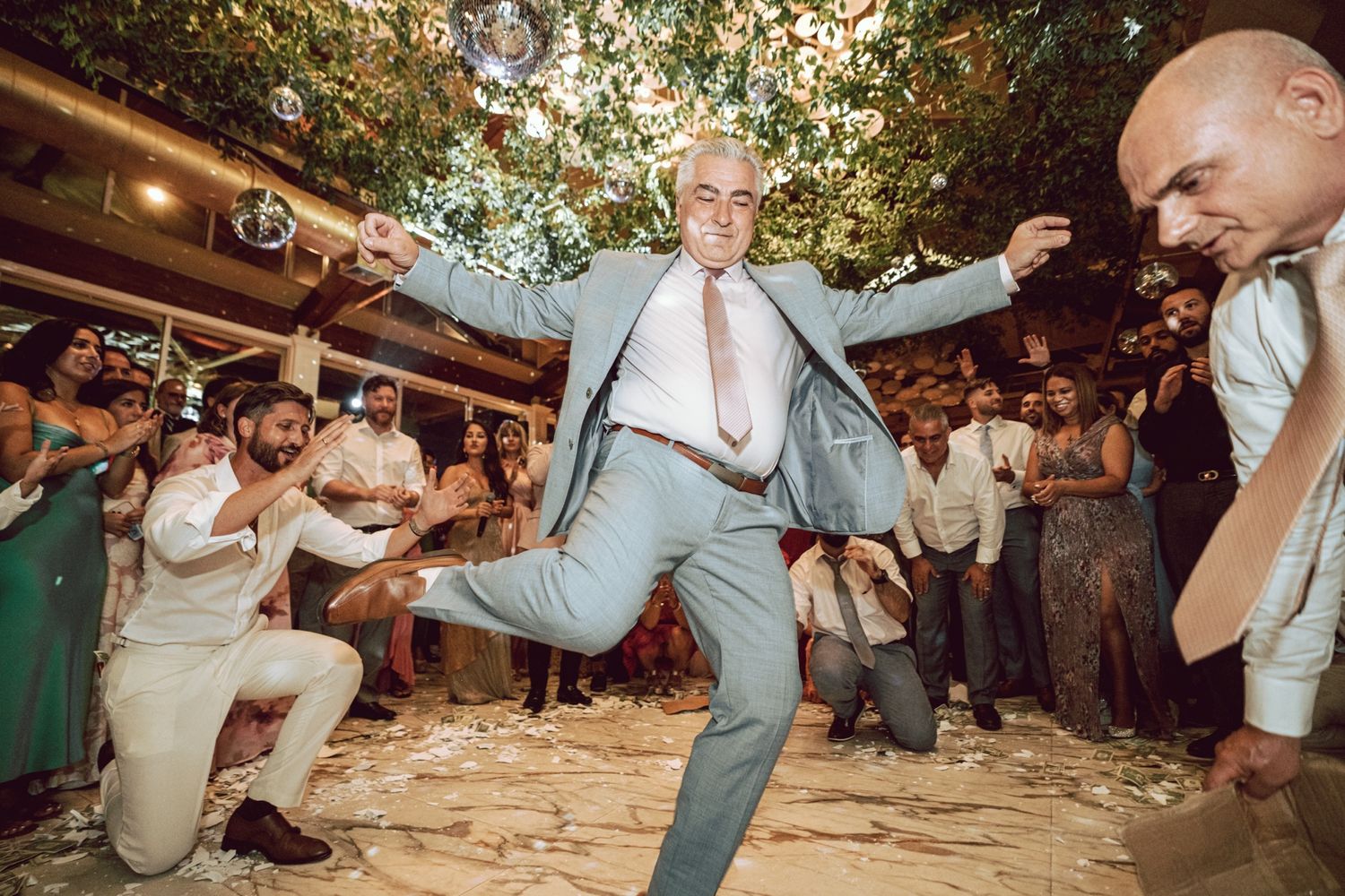 Man in light blue suit dancing exuberantly at outdoor wedding reception with guests.