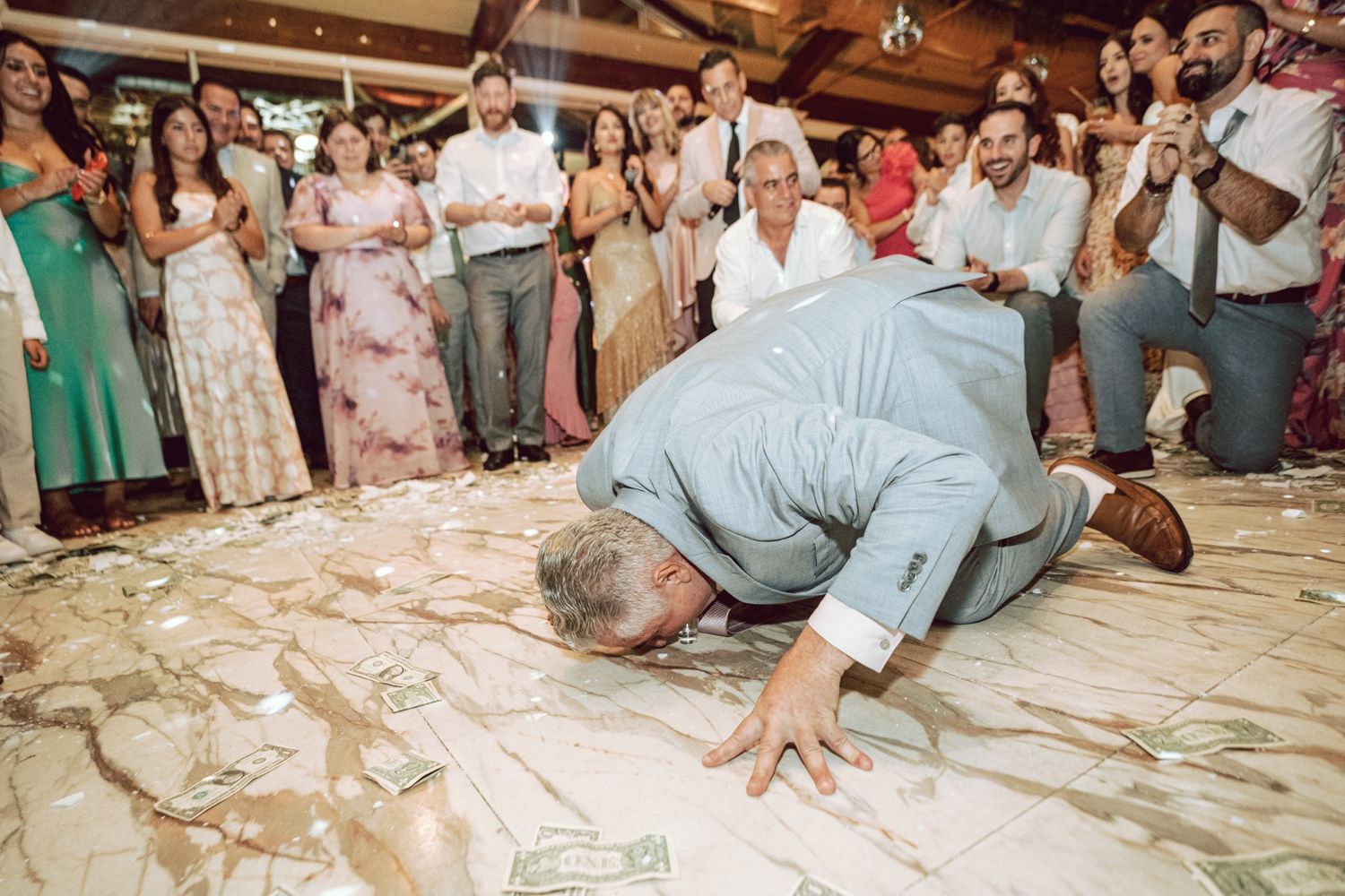 Man kneeling on a dance floor covered in money bills during a festive wedding reception celebration.